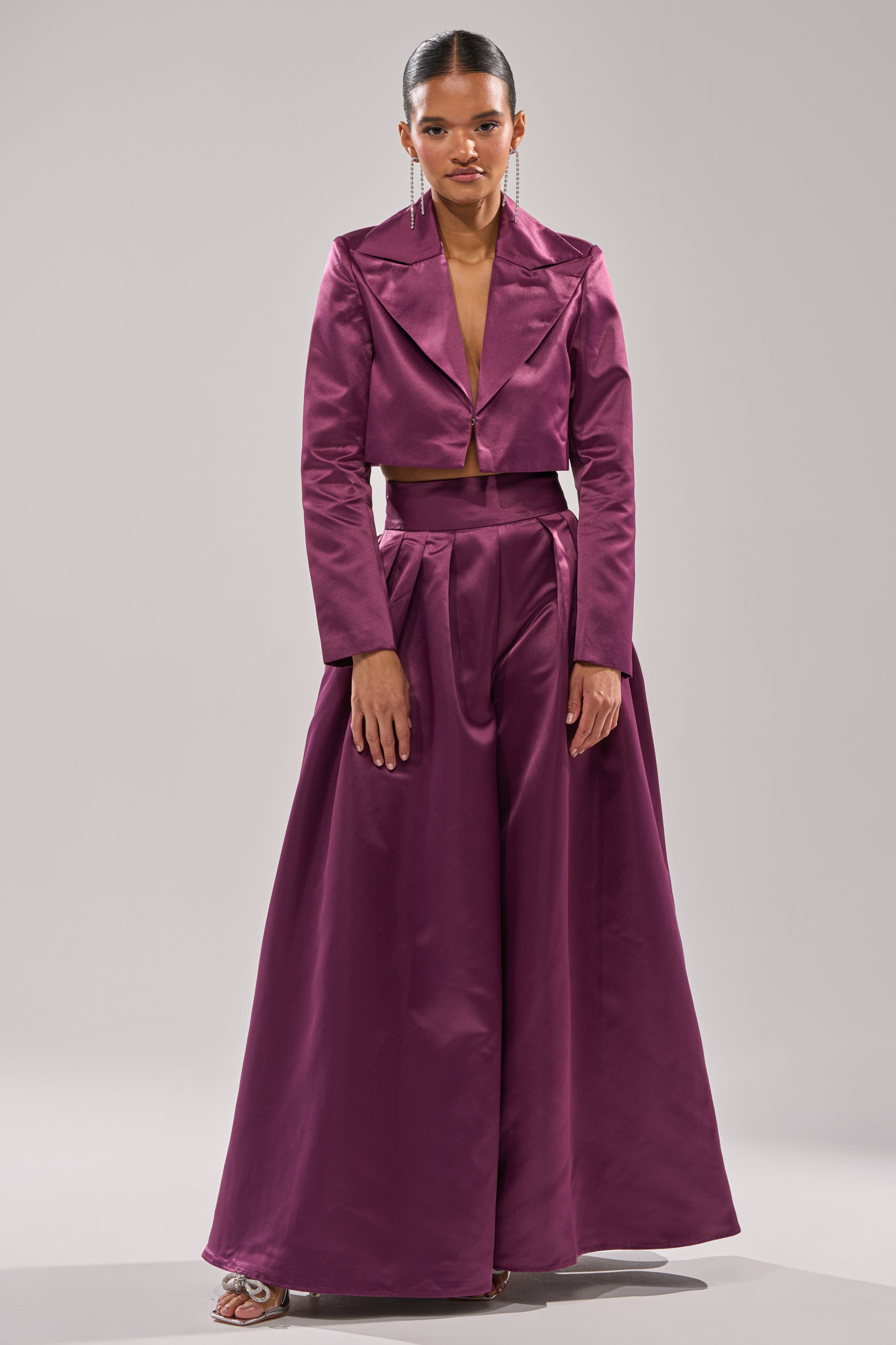 Wearing the ON AND ON CROPPED SATIN BLAZER and matching high-waisted, wide-leg pants in deep magenta, a woman poses against a plain background. Slicked-back hair and dangling earrings finish her elegant look.