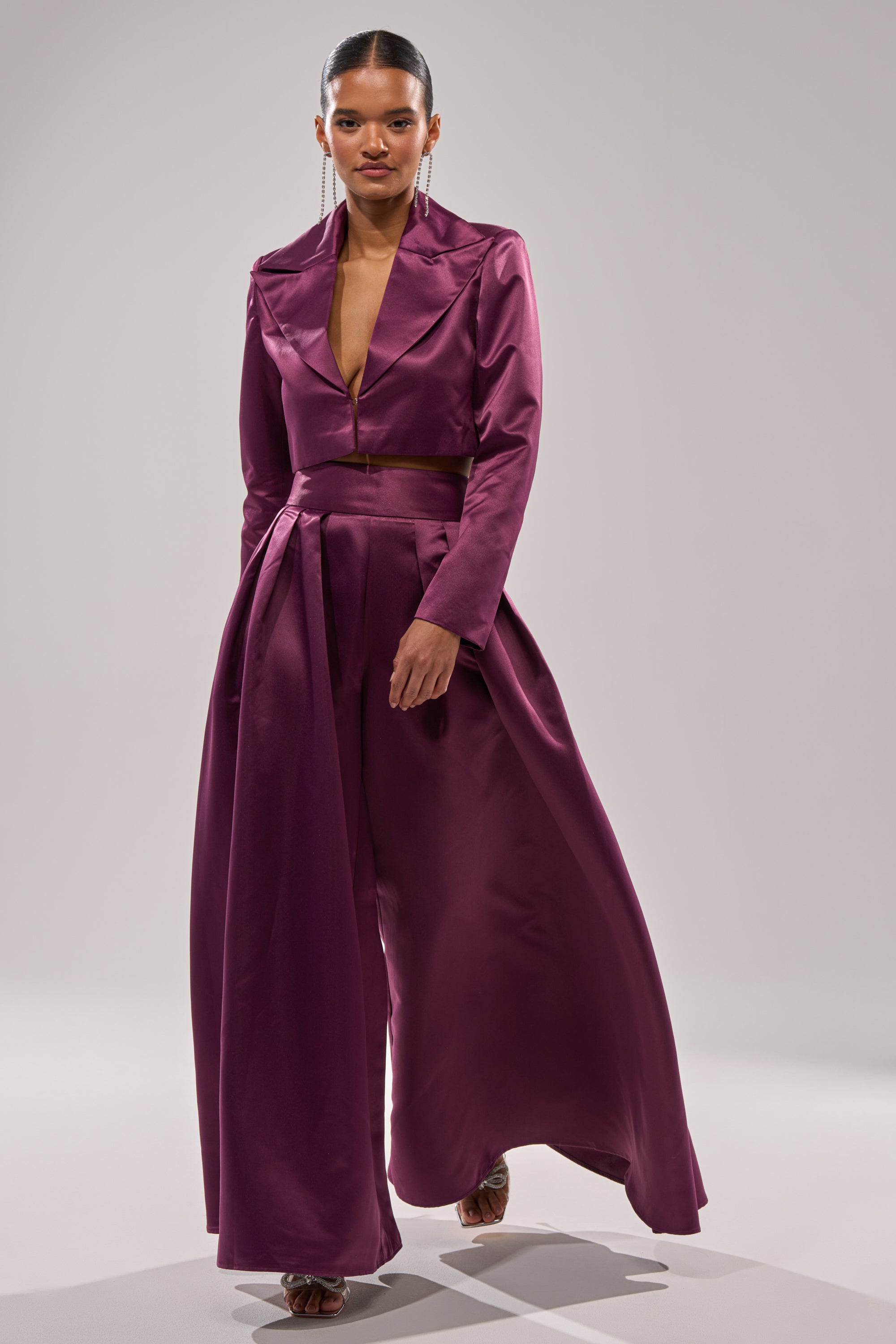A woman stands confidently against a plain gray background, wearing the ON AND ON CROPPED SATIN BLAZER styled with wide-legged satin pants.