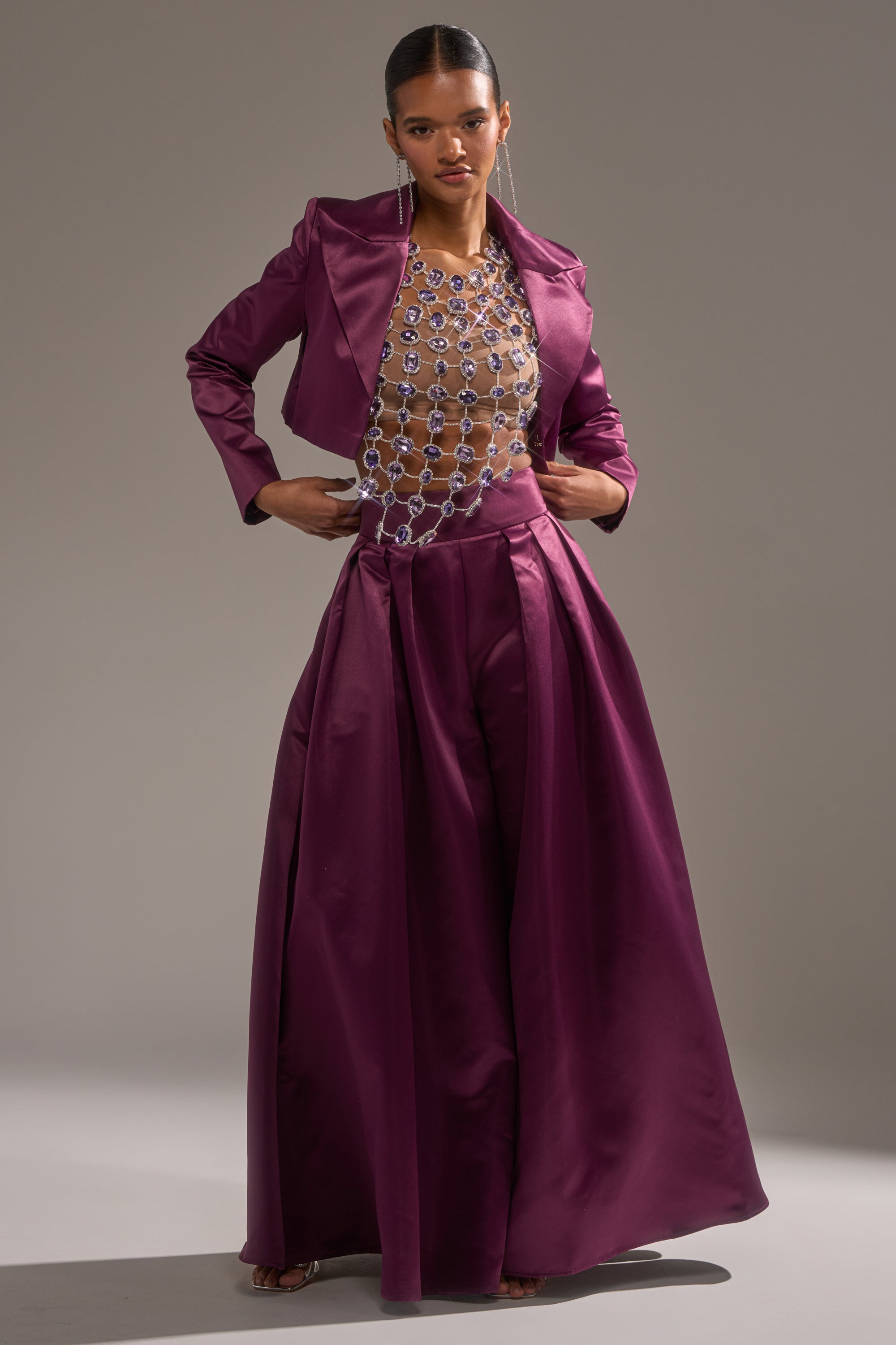 A woman exudes confidence in the ON AND ON CROPPED SATIN BLAZER—a shiny purple piece—styled with wide-leg pants and a sheer top with bold silver chain-link details, set against a simple neutral backdrop.