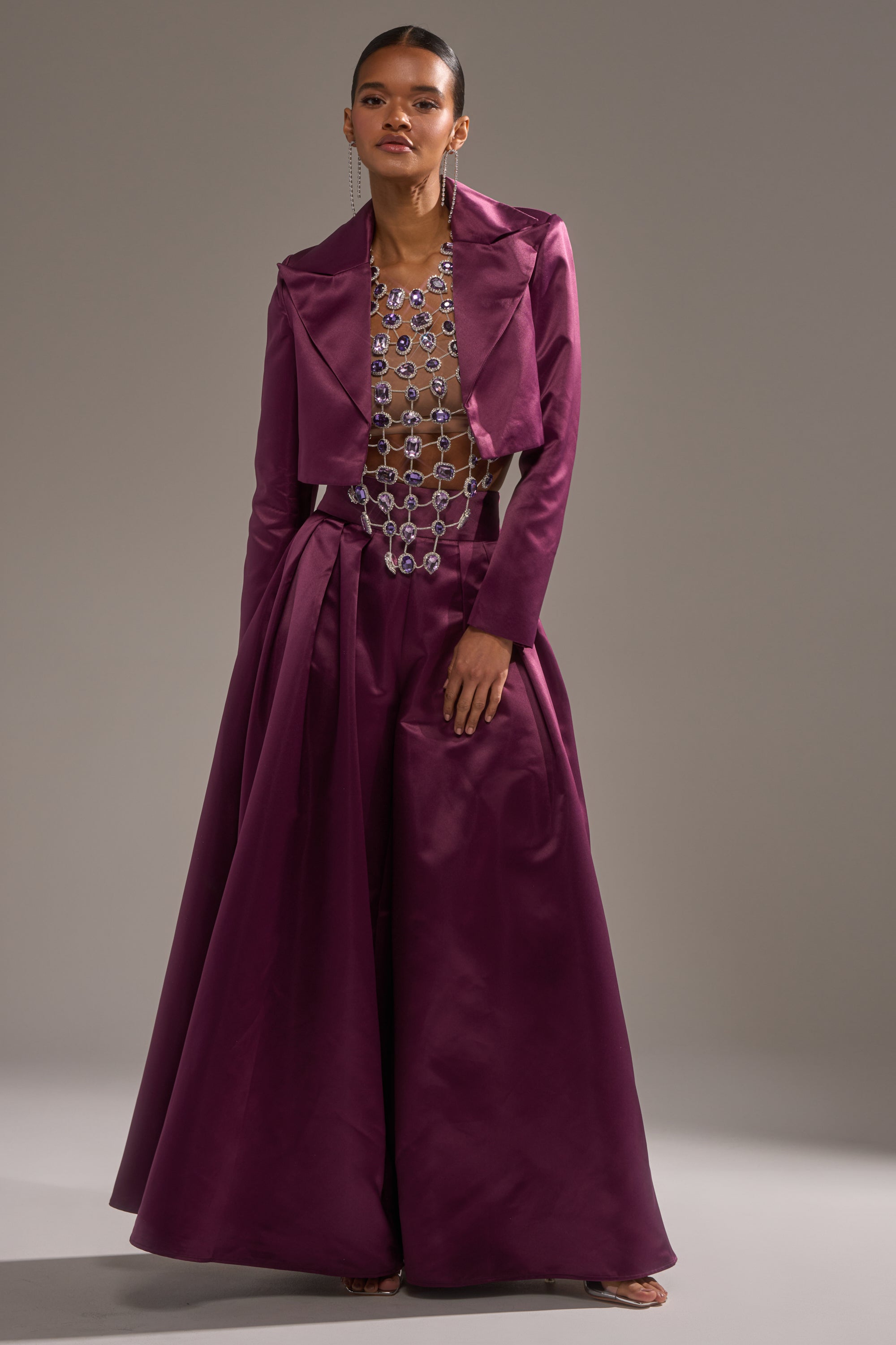 A woman poses against a neutral background wearing the ON AND ON CROPPED SATIN BLAZER in eggplant purple with wide-leg trousers and a sheer, silver-embellished top—a chic and modern ensemble.