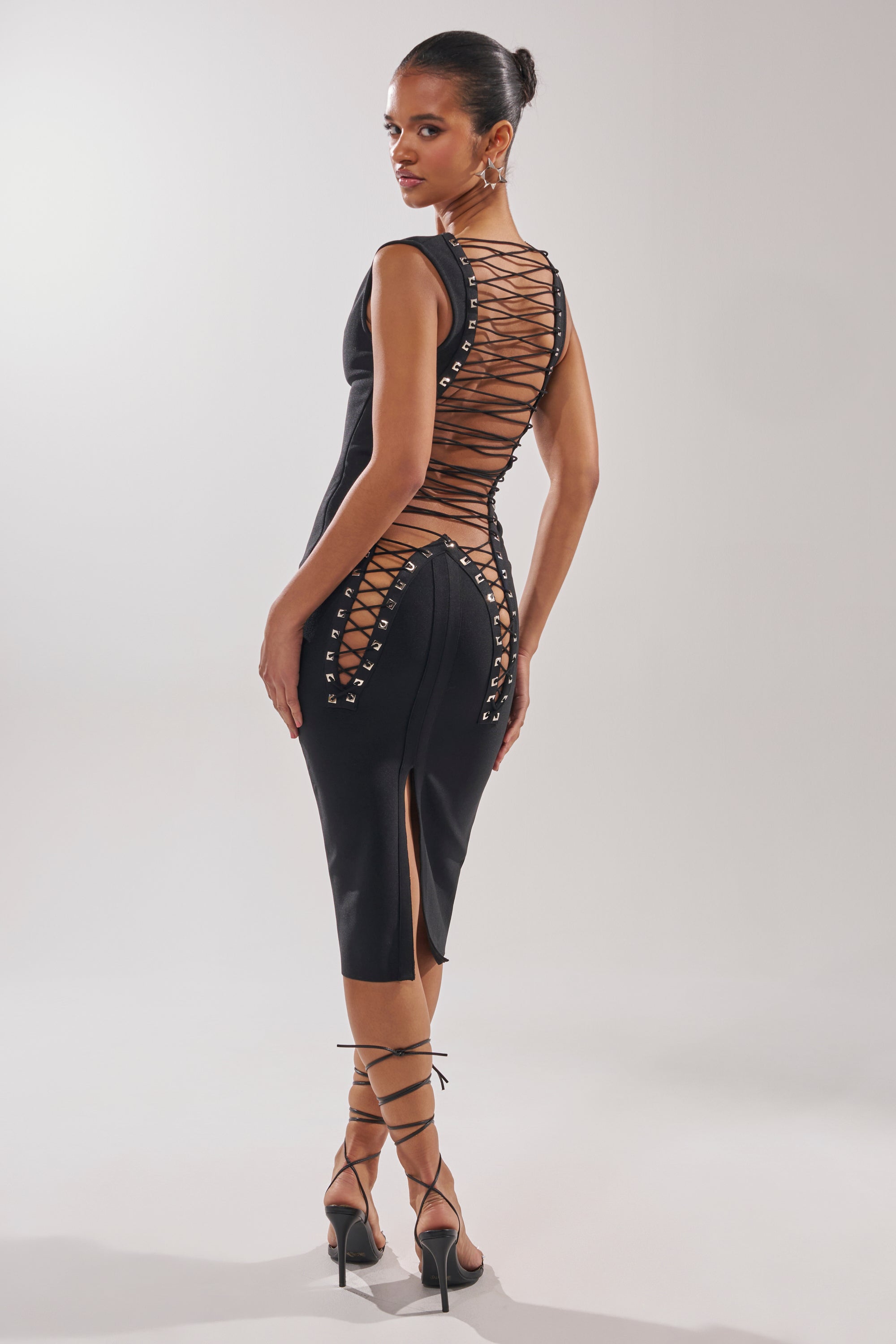 A woman models the MIDNIGHT LACES CORSET MIDI DRESS, showcasing sleek stretch bandage fabric with lace-up panels on the back and sides. She pairs it with strappy heels and a chic updo, posing against a plain light background.