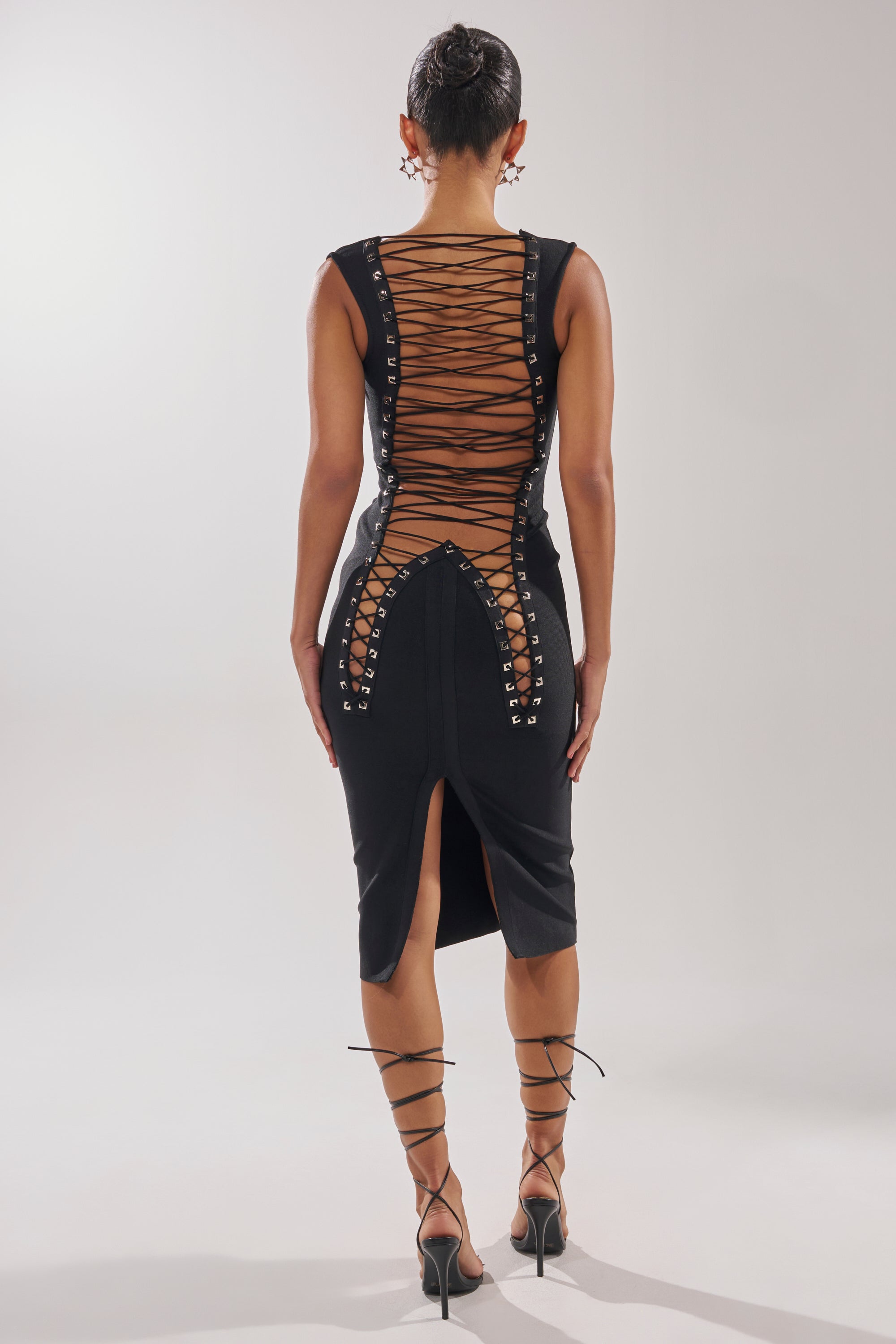 A woman stands facing away in the MIDNIGHT LACES CORSET MIDI DRESS—a fitted black lace-up back dress with crisscross details—paired with strappy heels and a sleek bun, set against a plain, light background.