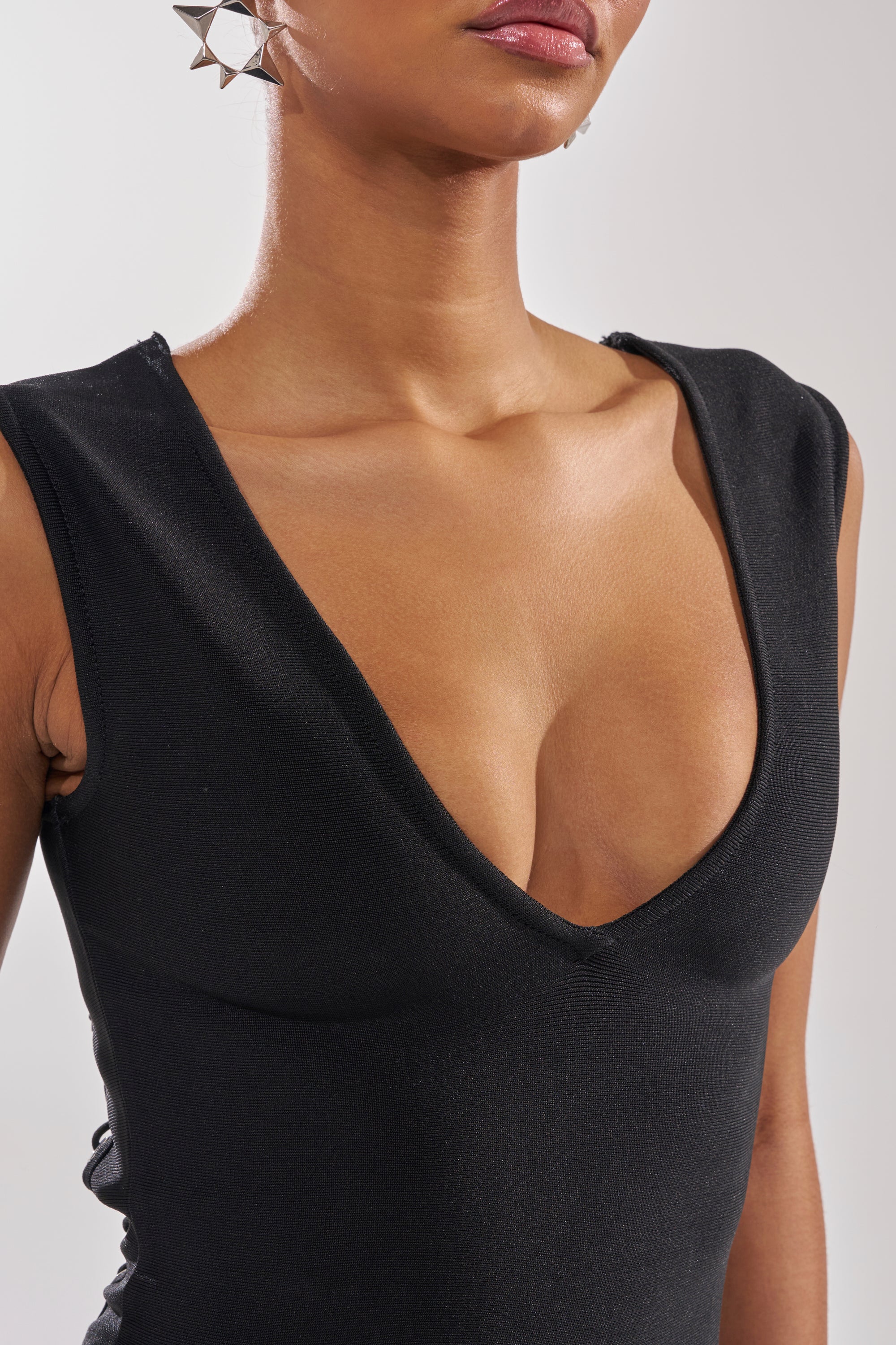 A person wearing the MIDNIGHT LACES CORSET MIDI DRESS—a sleeveless, deep V-neck black bandage fabric dress—paired with large, spiked silver earrings, shown from shoulders to chest against a plain background.