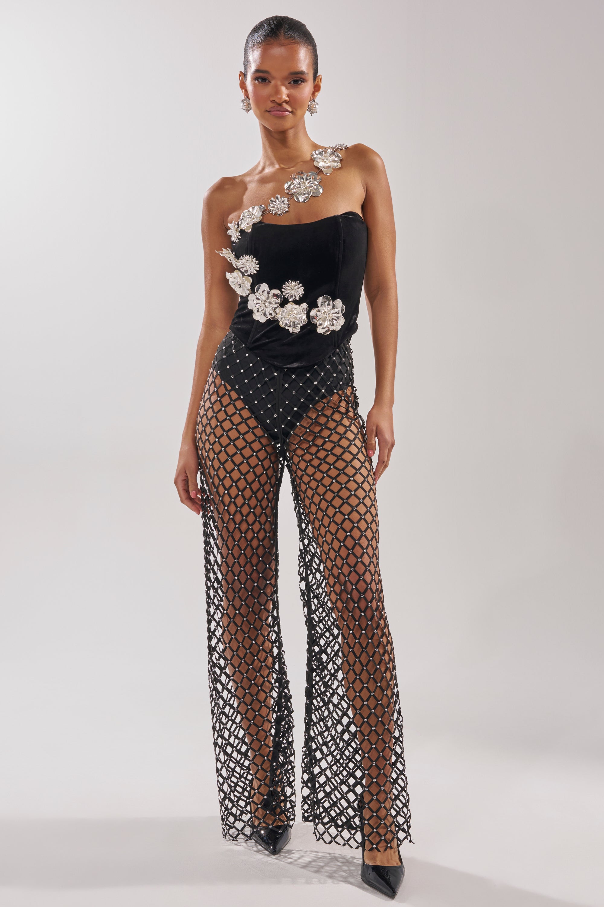 A woman stands confidently against a plain background wearing CELINE STUD MESH PANT styled with a black, one-shoulder bodysuit featuring large white floral embellishments and black high heels. 0