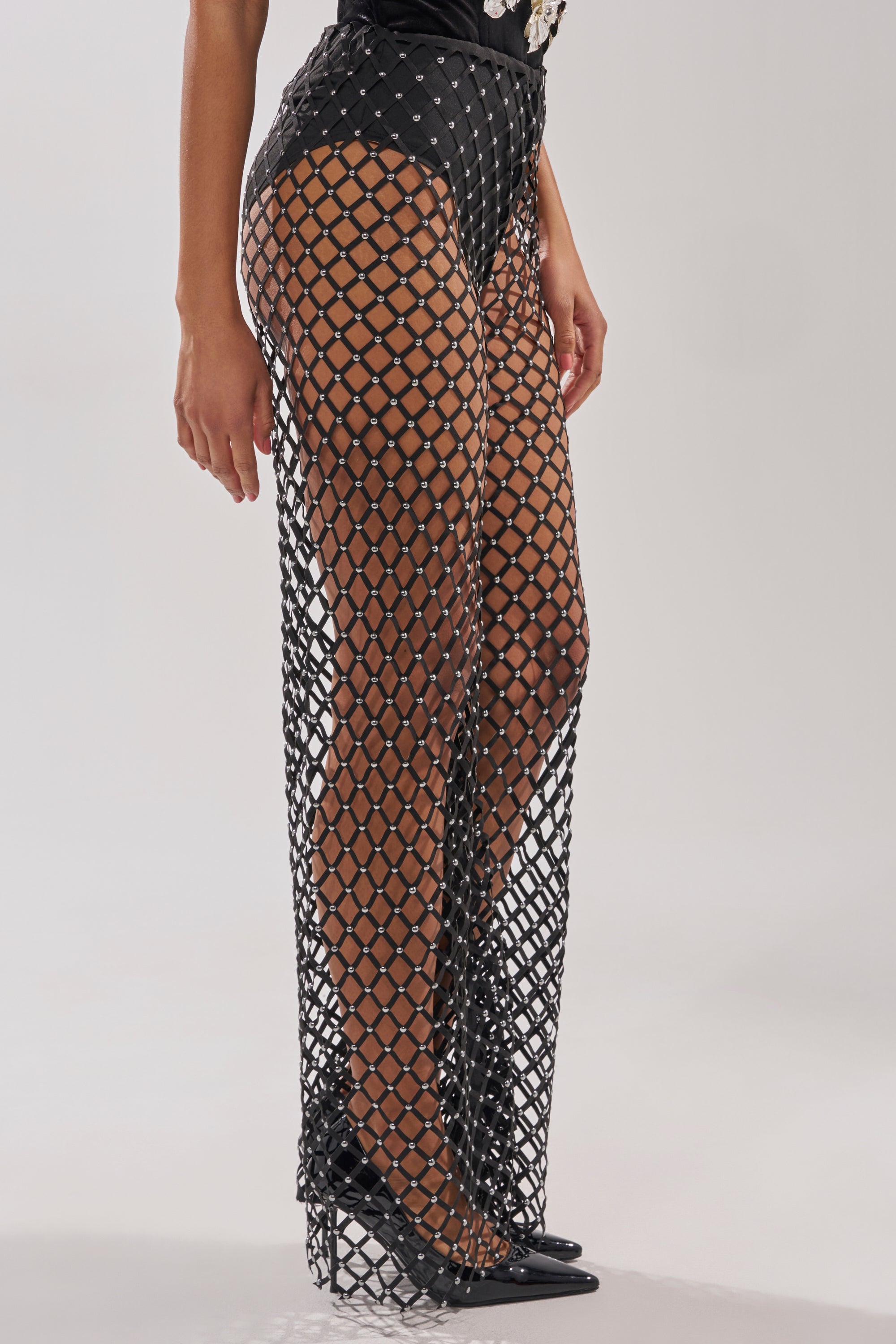 A person wears CELINE STUD MESH PANT—black, high-waisted bottoms with a large diamond-shaped net pattern—over black underwear, paired with black pointed-toe heels. 3