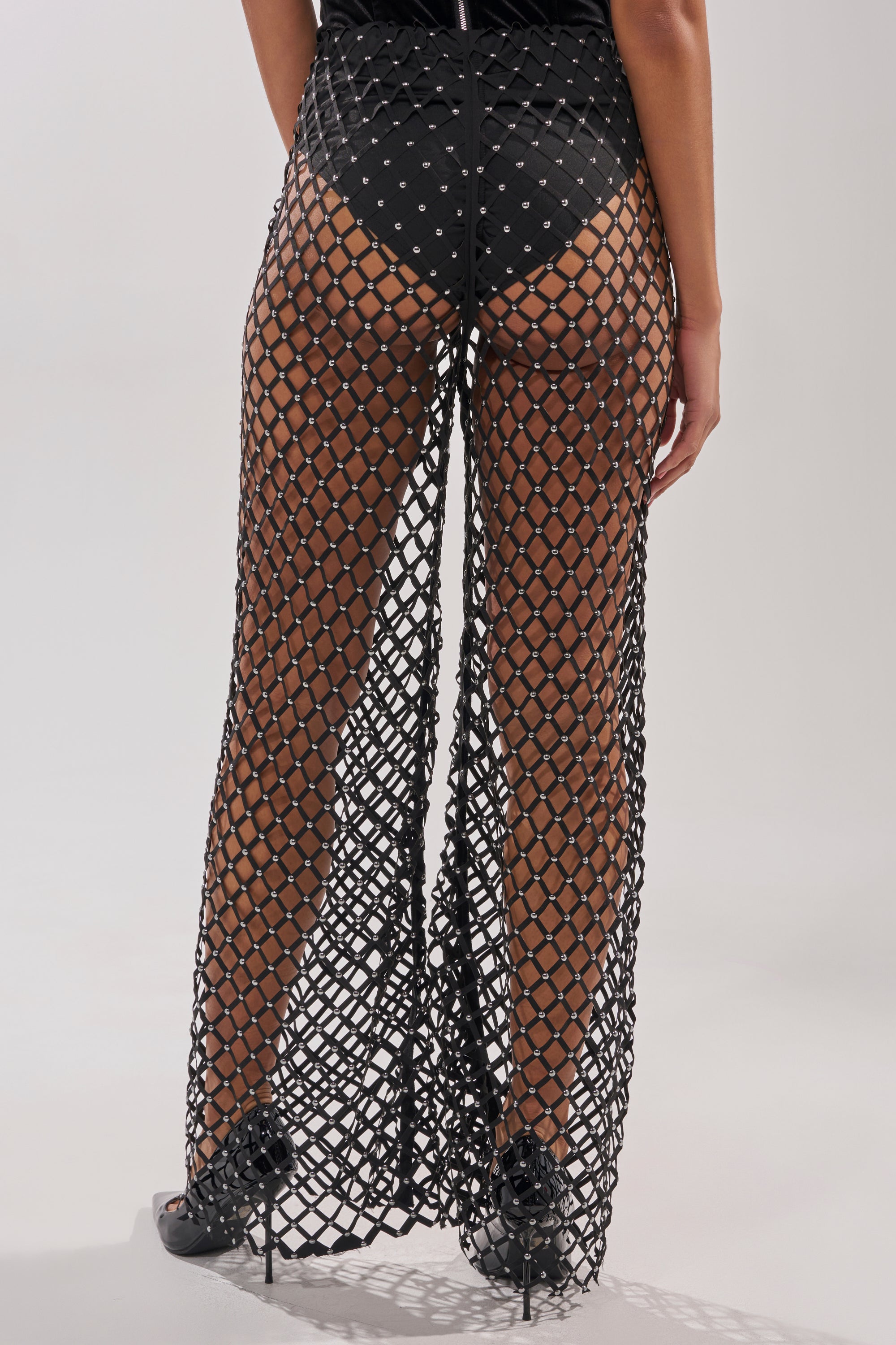 A person, seen from behind, wears the CELINE STUD MESH PANT—sheer, floor-length mesh with large diamond-shaped openings over black underwear—and black high-heeled shoes. 4