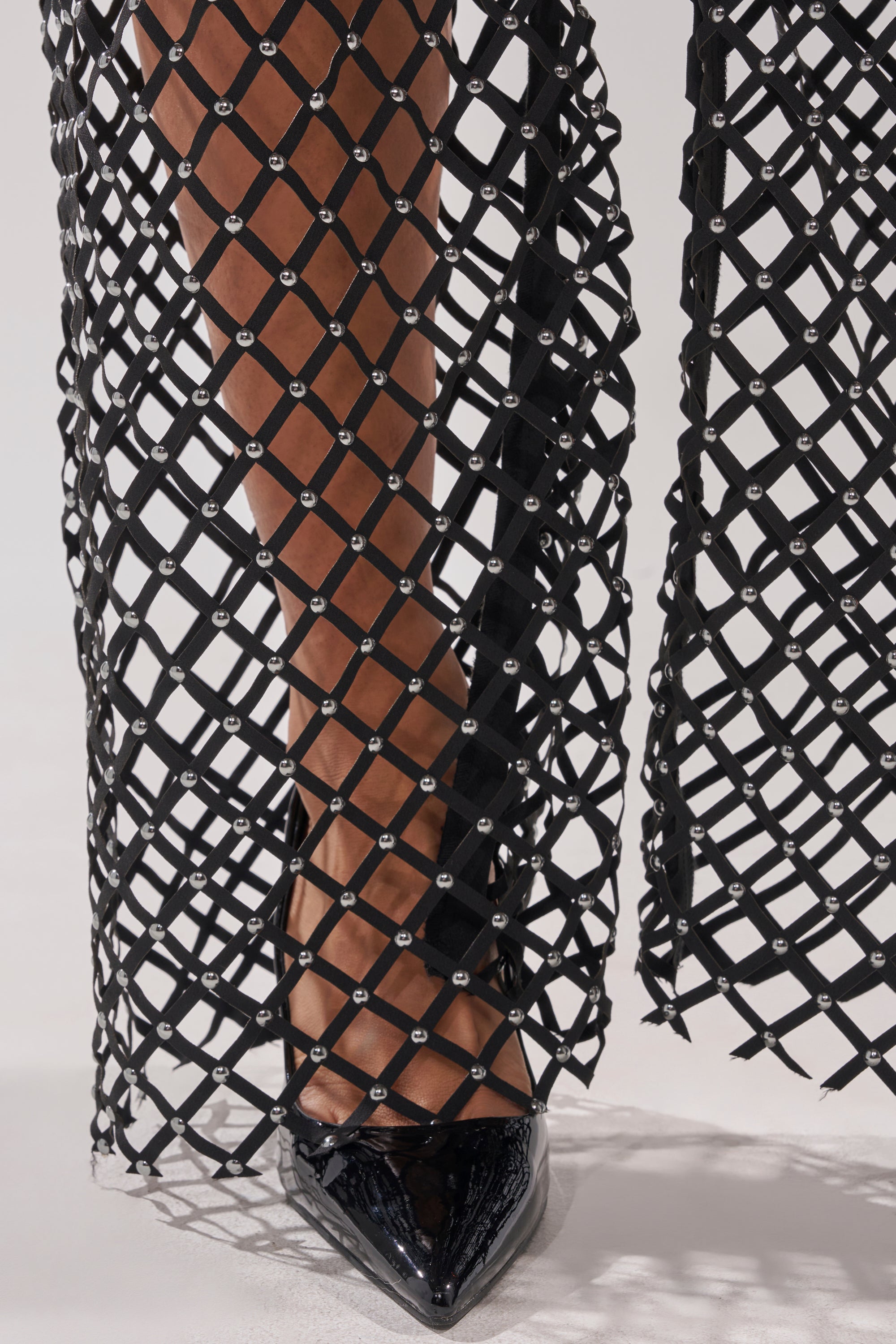 A close-up of a person’s lower leg wearing the CELINE STUD MESH PANT with rhinestone embellishments and black pointed-toe patent leather heels, standing on a light-colored surface. 8