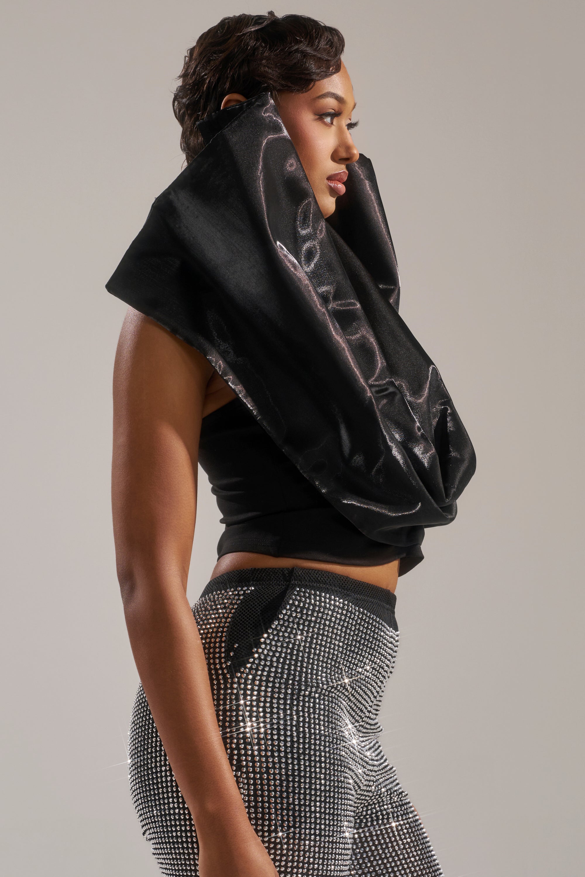 A woman in profile wears the DROP IT LOW BLOUSE—a sculptural satin top with a large black collar—paired with high-waisted, silver sparkling pants. Its cropped fit adds modern edge against a plain, neutral background. 3