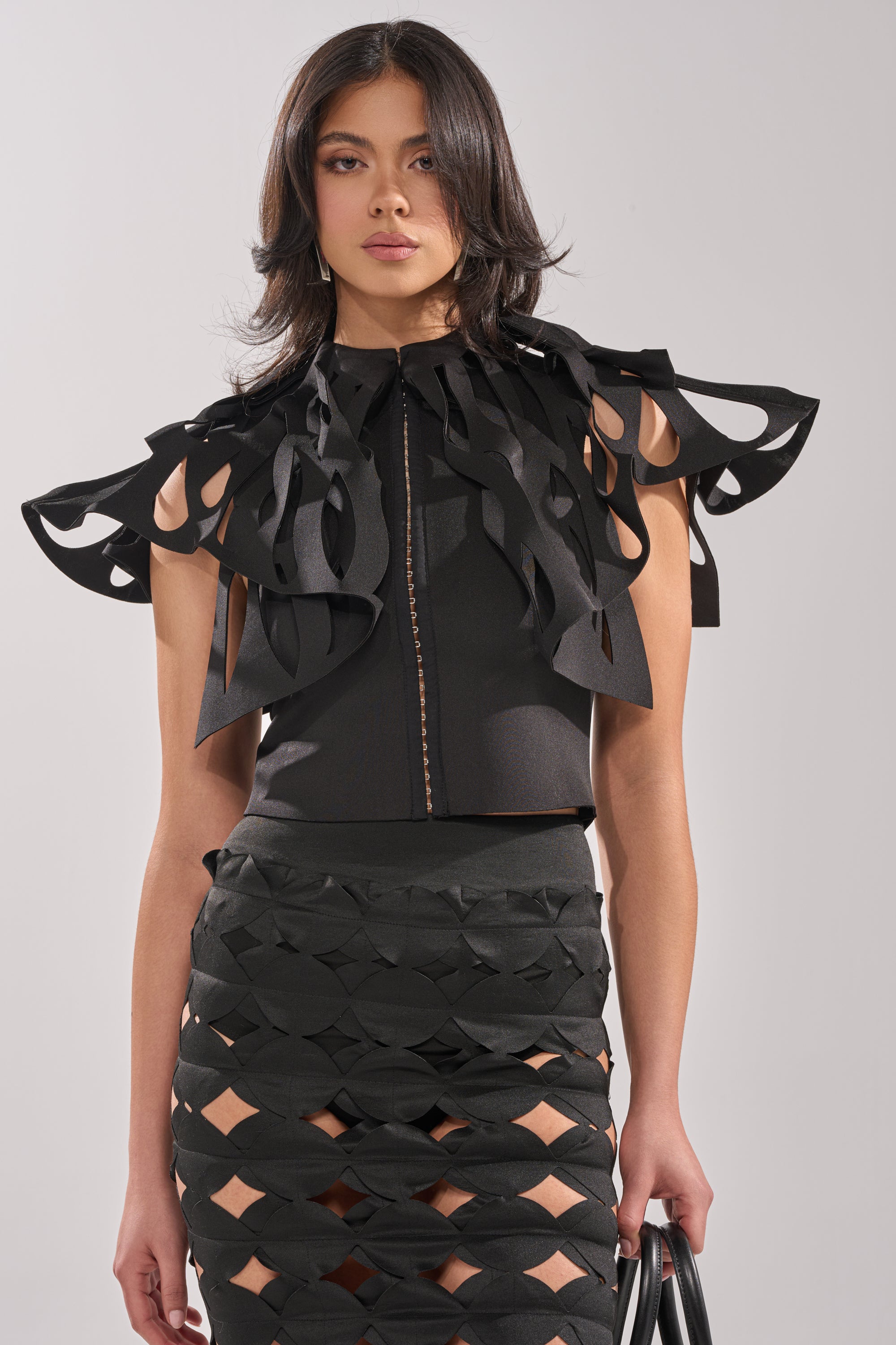 A woman wears the SEGA BLOUSE IN BLACK with a matching skirt featuring intricate cut-out patterns as she stands against a plain background. 1