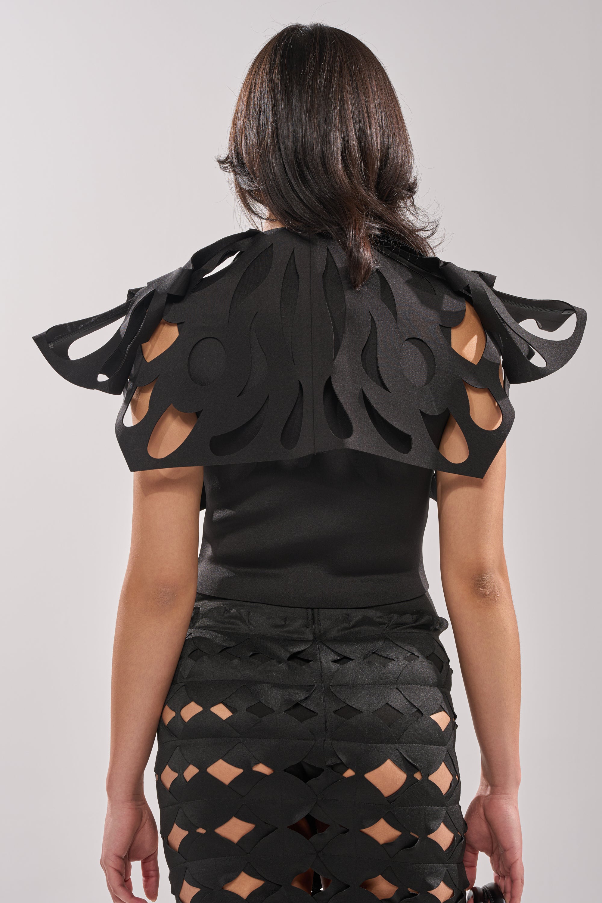 A woman faces away, wearing the SEGA BLOUSE IN BLACK featuring sculptural cut-out details on the shoulders and skirt, exposing skin through geometric openings. Her dark hair flows in loose waves against a plain light gray background. 3