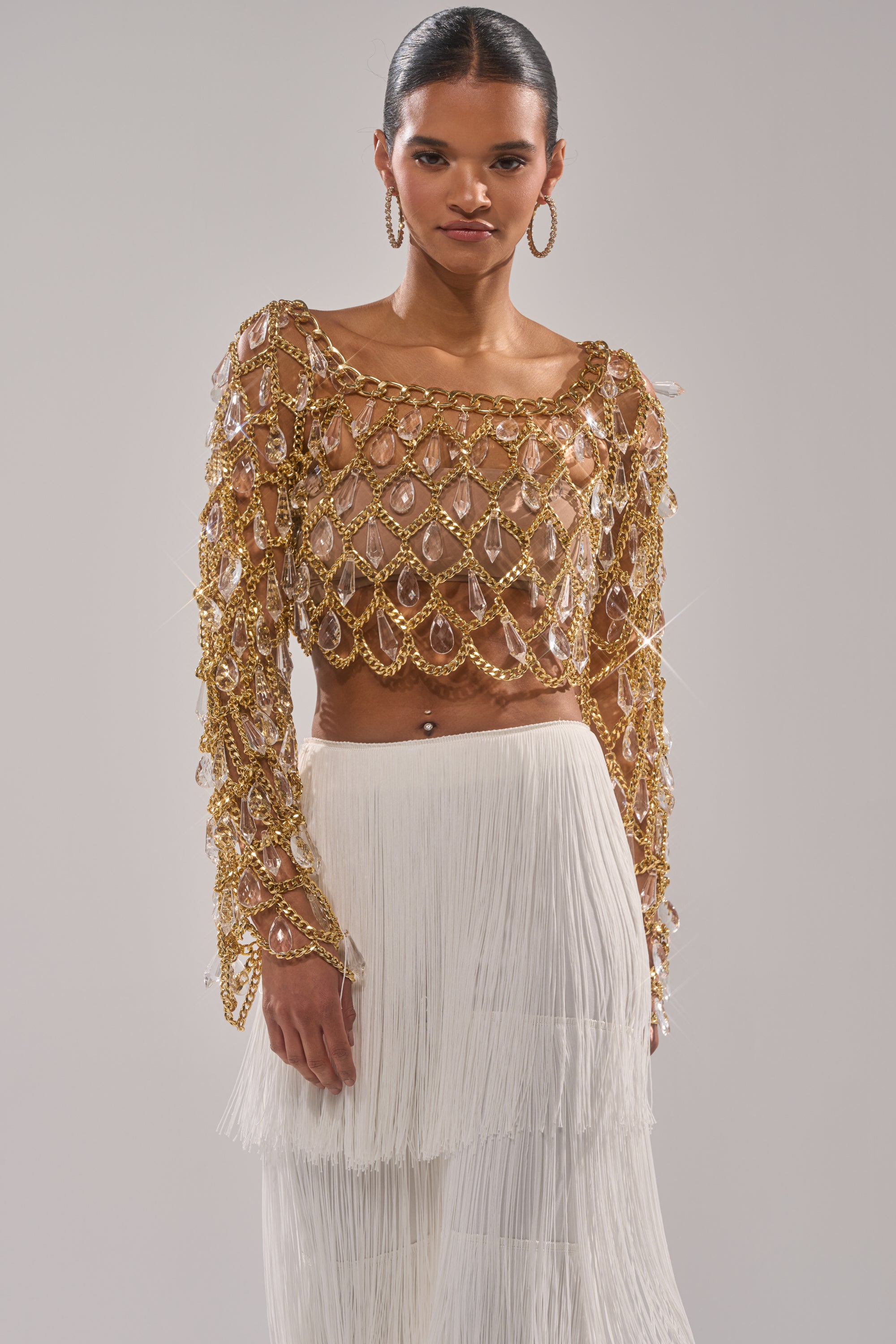 Wearing the GLAM BABY CHAIN TOP with crystal embellishments over a nude underlayer, paired with a white fringe-layered skirt, a person stands against a plain light gray background. 0