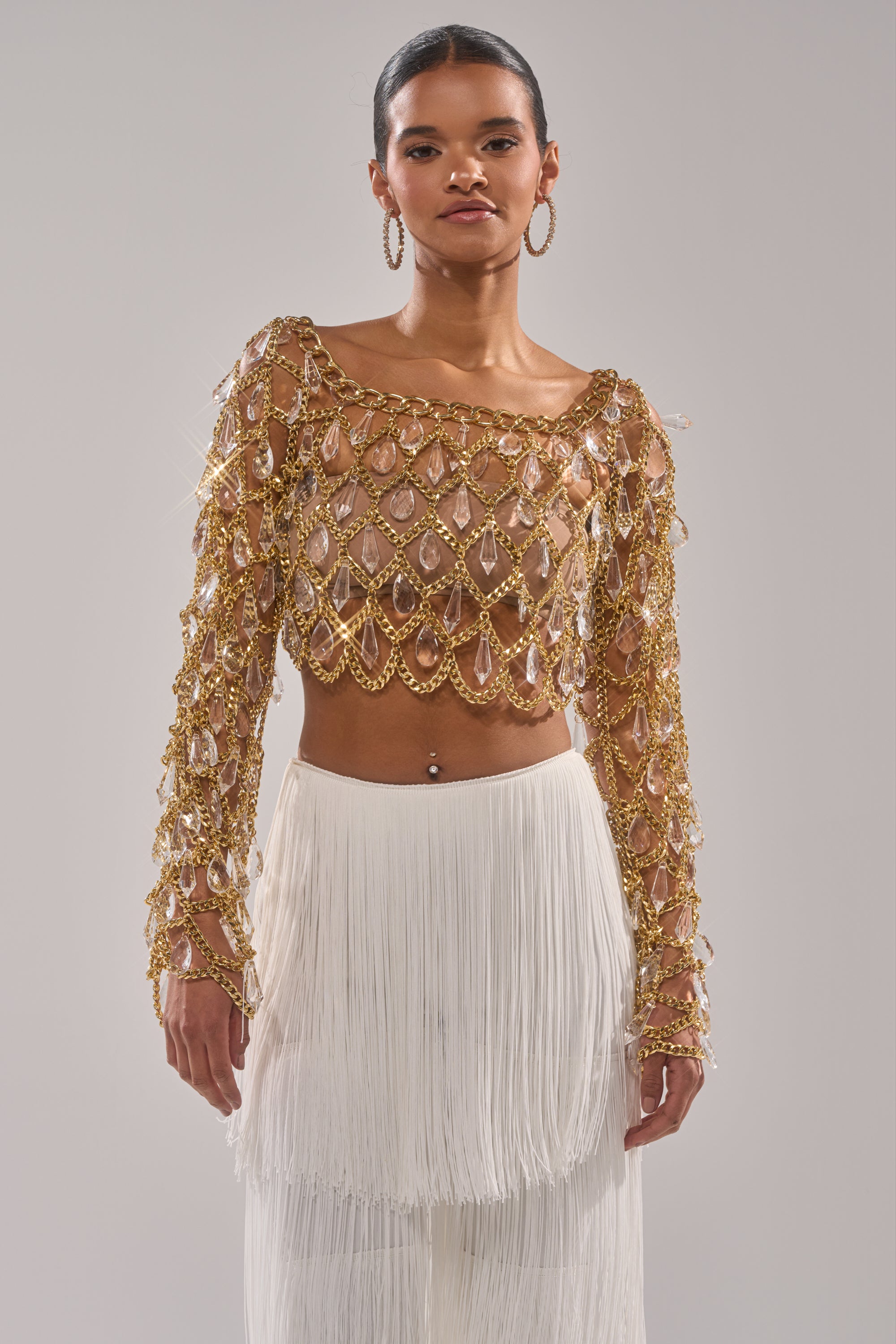 A woman stands facing forward in the GLAM BABY CHAIN TOP, a gold chain mesh top with teardrop details over sheer fabric, paired with a high-waisted white fringed skirt. She wears hoop earrings, has sleek hair, and sports a navel piercing. 1