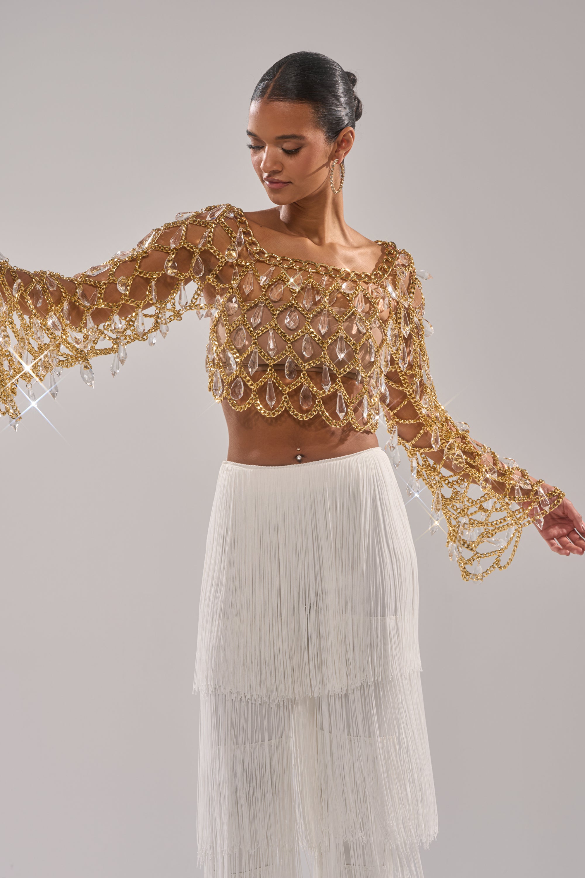 A woman models the GLAM BABY CHAIN TOP—a gold, crystal-embellished design with bell sleeves—paired with a white fringed skirt, striking a graceful pose as the sparkling details catch the light. 2