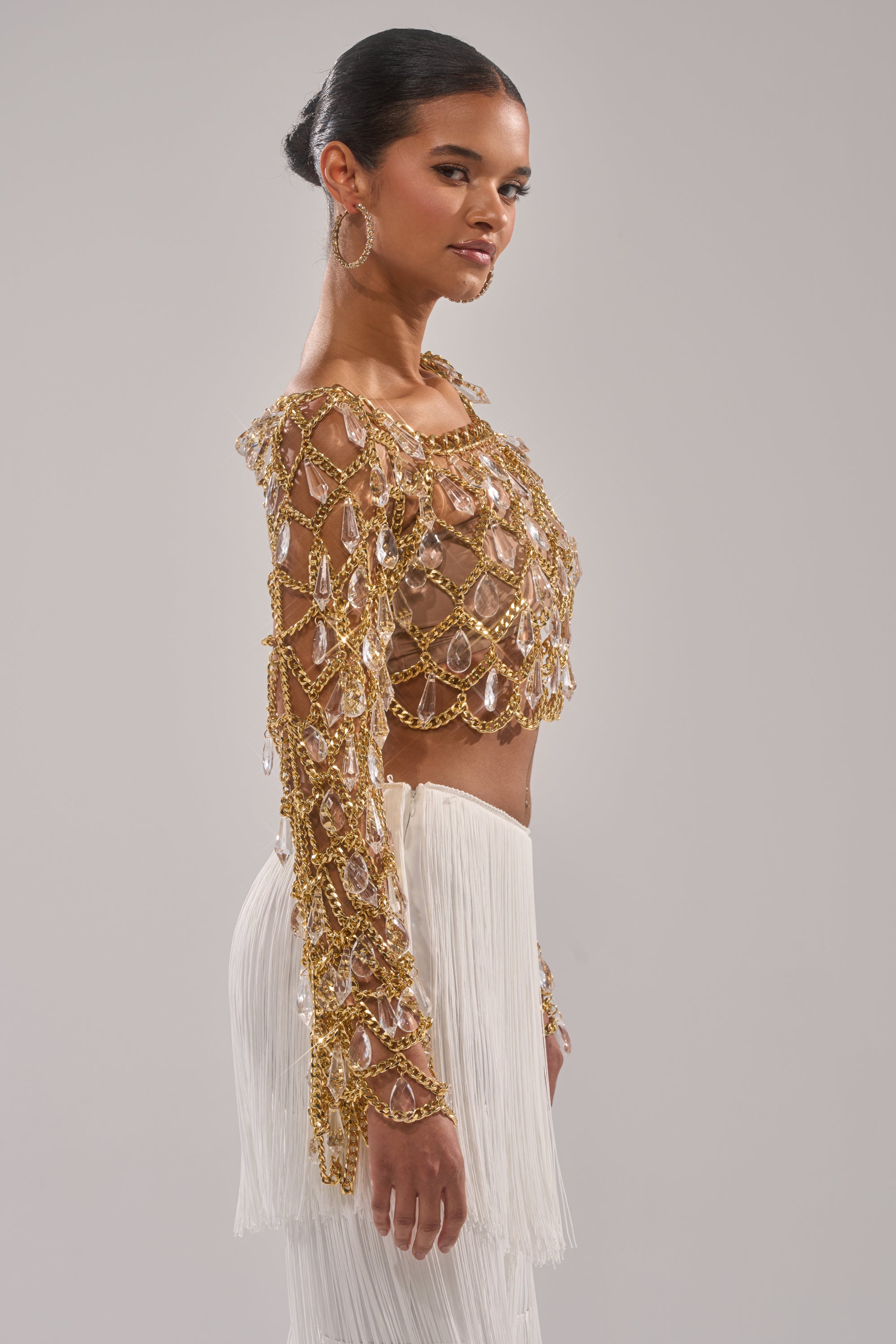 A woman in profile wears the GLAM BABY CHAIN TOP with crystals, gold chains, and gems, paired with a white fringed skirt. Her sleek bun and large hoop earrings complete the look against a plain background. 3