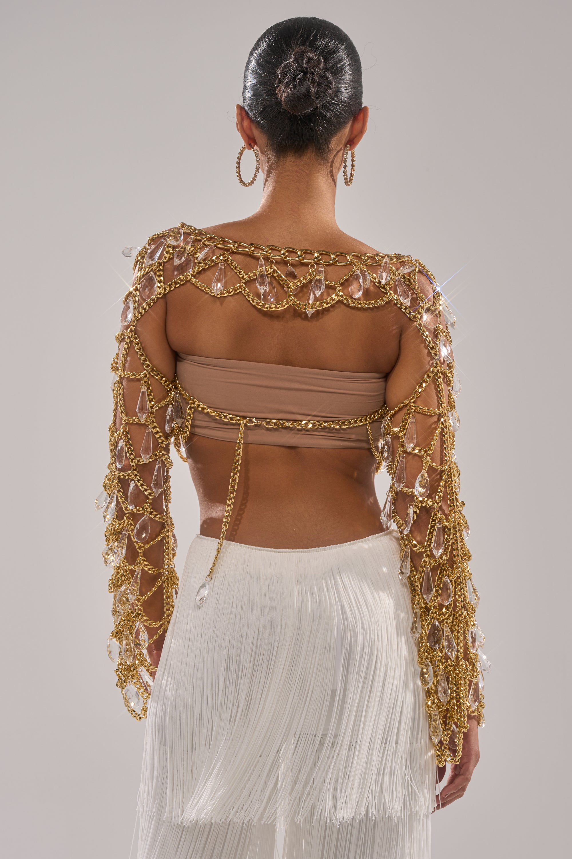 A woman with her hair in a slicked-back bun and large hoop earrings wears the GLAM BABY CHAIN TOP, which features gold-tone chains and crystal embellishments over a tan bandeau, paired with a white fringed skirt. 4