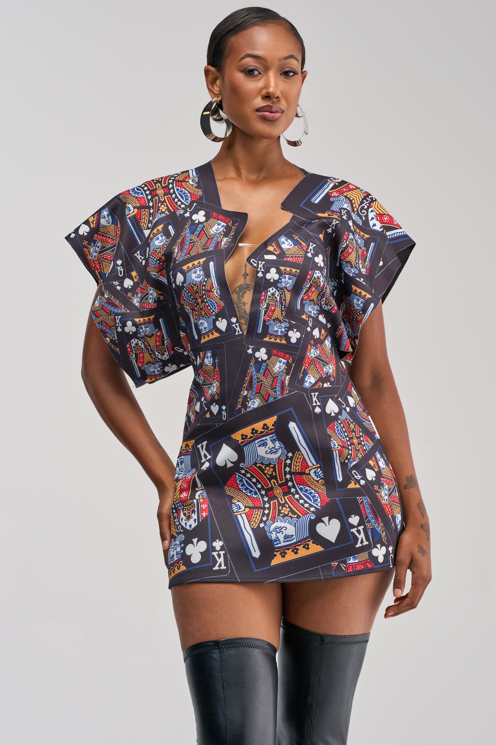 A woman poses confidently in the HOUSE OF CARDS MINI DRESS IN BLACK MULTI, which features a bold king card print, deep neckline, and wide sleeves. She accessorizes with hoop earrings and thigh-high black boots, standing against a plain light background.