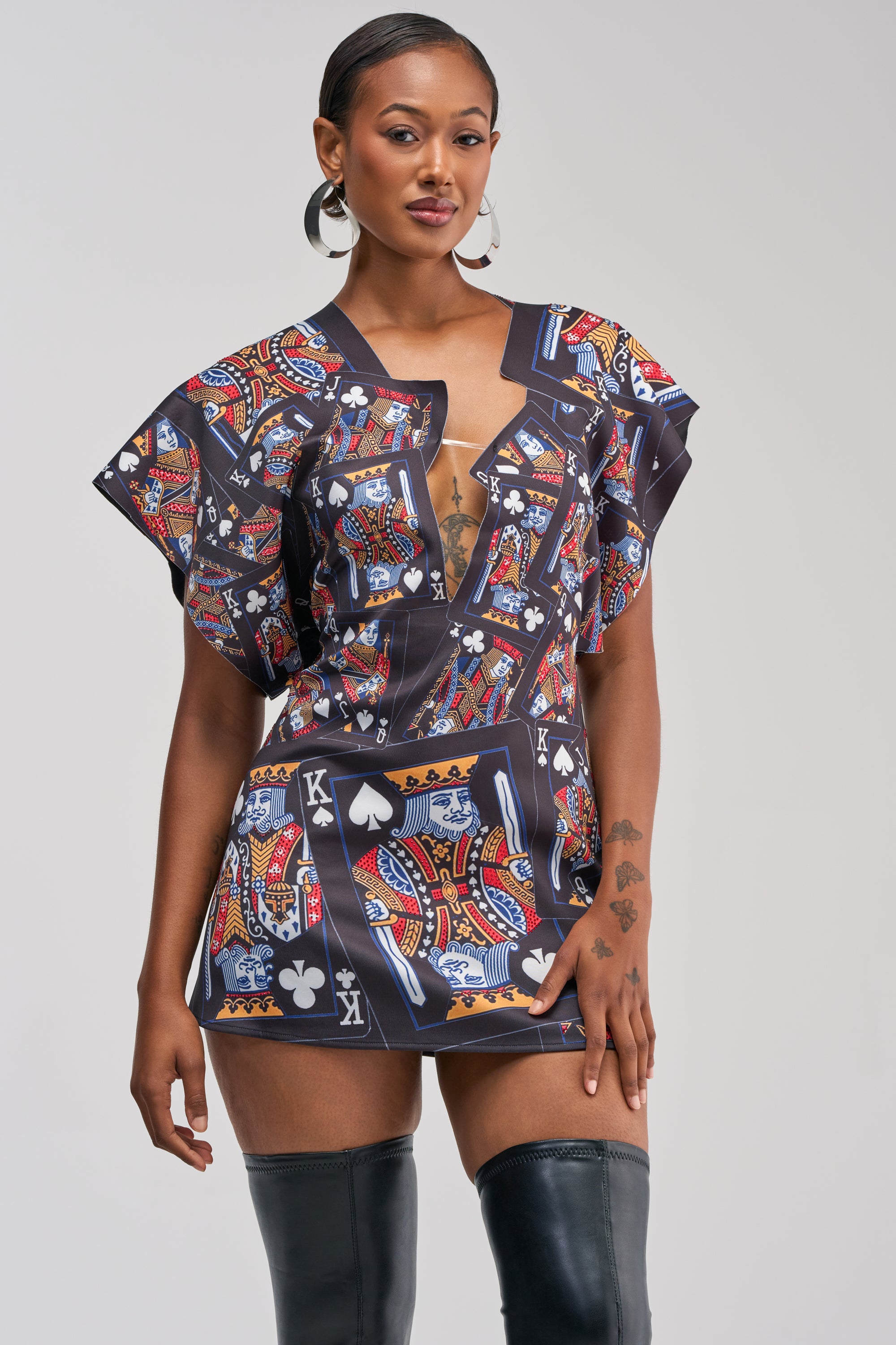 A woman stands confidently against a plain background, dressed in the HOUSE OF CARDS MINI DRESS IN BLACK MULTI, which features a striking playing card motif; she completes her look with large hoop earrings and over-the-knee black boots.