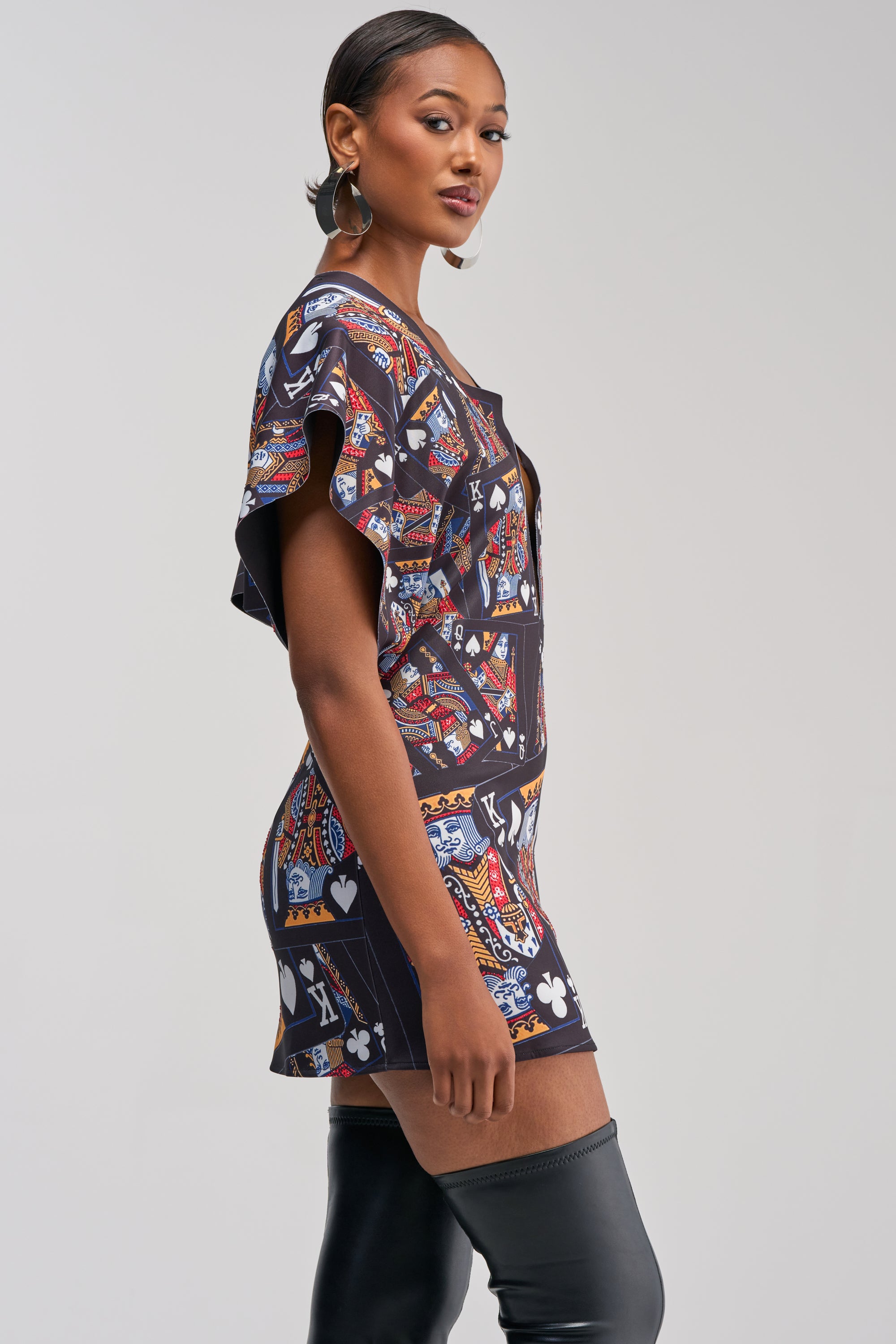 A woman stands sideways, striking a confident pose against a plain light background in the HOUSE OF CARDS MINI DRESS IN BLACK MULTI, featuring a vibrant king card print and flutter sleeves, paired with thigh-high black boots.