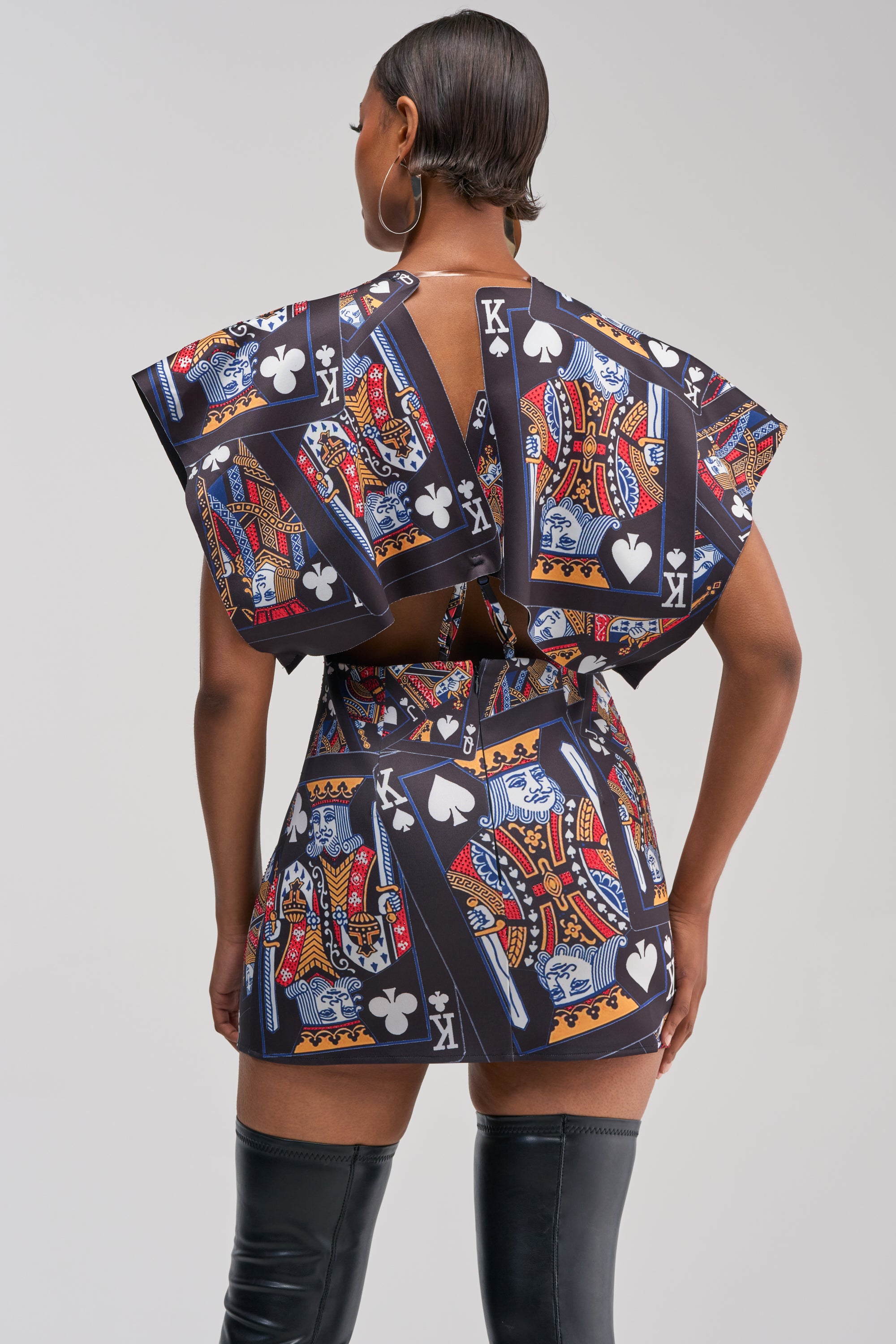 Wearing the HOUSE OF CARDS MINI DRESS IN BLACK MULTI, which features a bold playing card motif with the King of clubs and spades, a person is shown from the back. They have short hair, accessorized with large hoop earrings and thigh-high black boots.