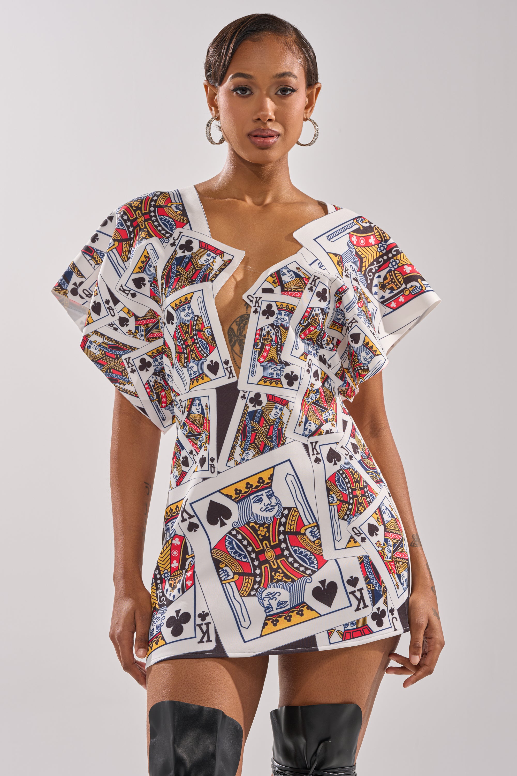 A woman stands confidently in the HOUSE OF CARDS MINI DRESS, a short dress with a playing card print, kings of various suits, and an asymmetrical plunge neckline, styled with hoop earrings and thigh-high black boots. 0