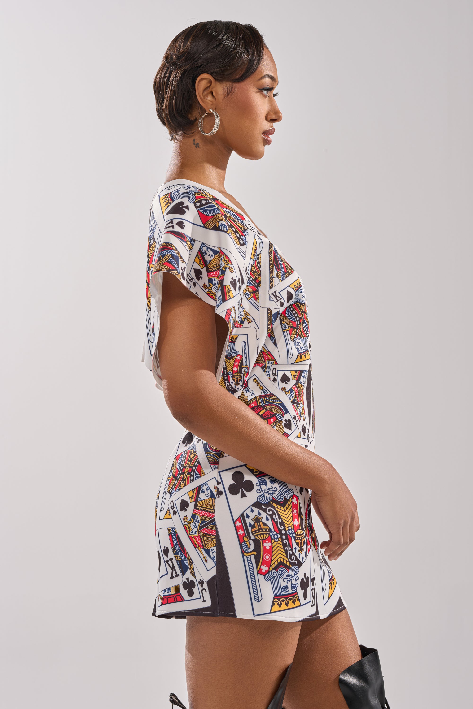 A woman in profile models the HOUSE OF CARDS MINI DRESS, featuring bold playing card prints, hoop earrings, and black boots against a plain backdrop. 2
