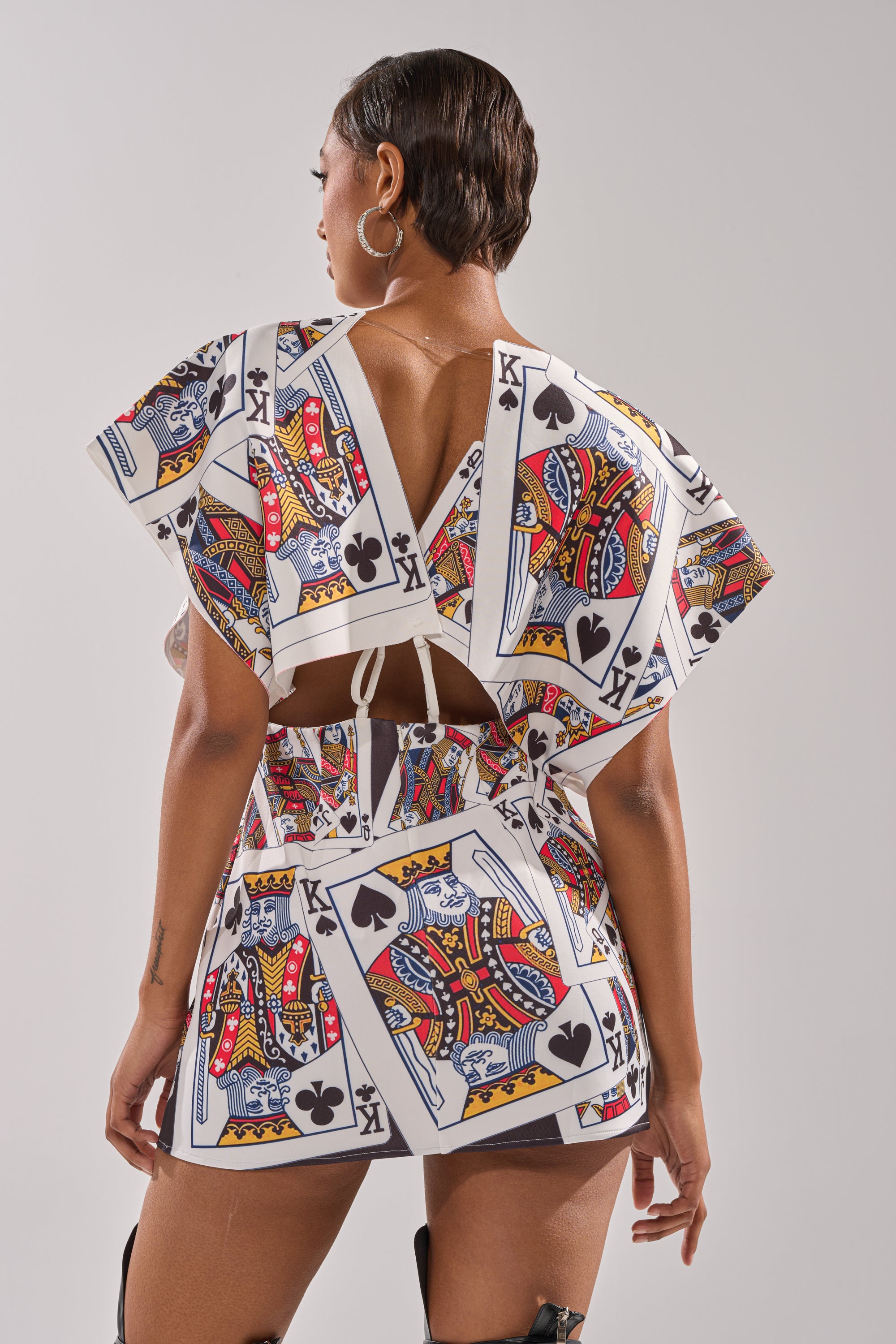A person stands facing away, wearing the HOUSE OF CARDS MINI DRESS—a bold matching top and skirt set featuring geometric shapes and oversized prints of the king of spades, clubs, and diamonds. 3