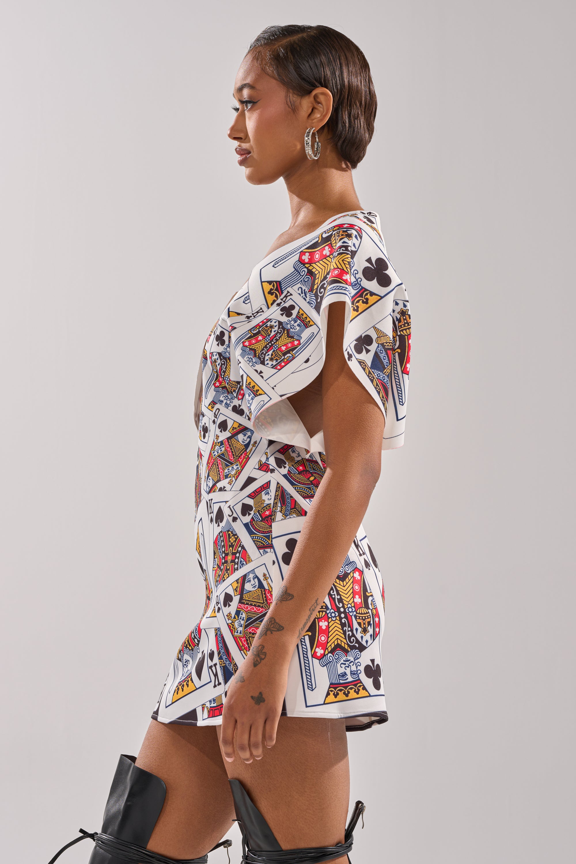A woman in profile models the HOUSE OF CARDS MINI DRESS, featuring a playing card print with queen and club motifs. She wears hoop earrings, has slicked-back hair, black knee-high boots, and stands against a plain light background. 4