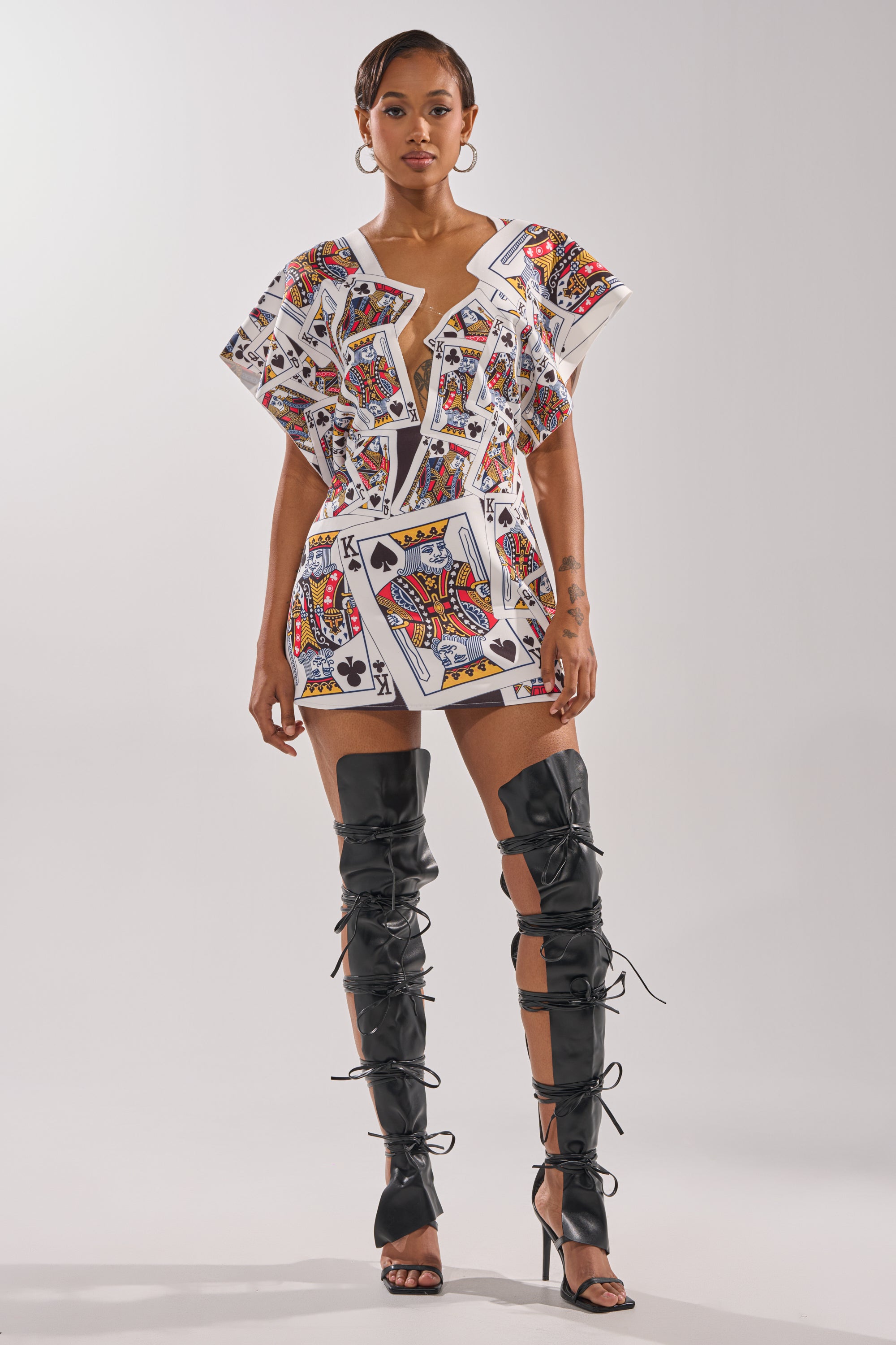 A woman stands confidently in the HOUSE OF CARDS MINI DRESS, a playing card print mini dress with an asymmetrical plunge neckline featuring the Queen of Spades, paired with thigh-high black lace-up boots against a white background. 5