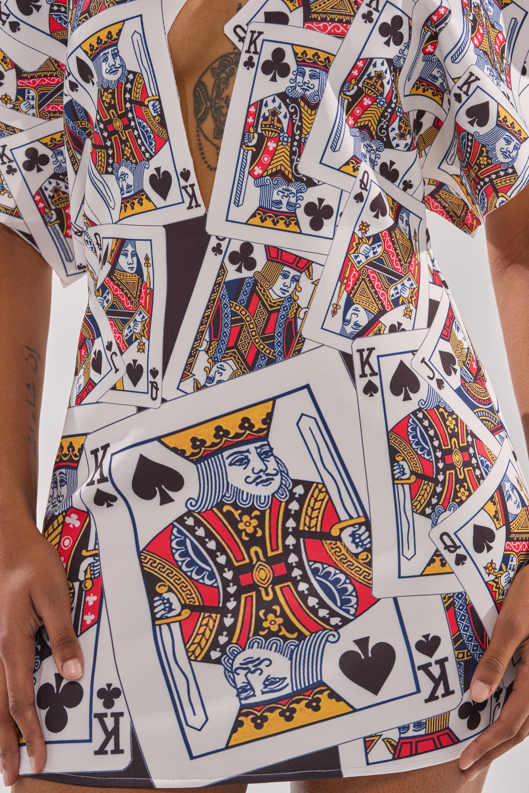 Wearing the HOUSE OF CARDS MINI DRESS, which features a playful playing card print with vibrant kings of spades and clubs, an asymmetrical plunge neckline, plus visible tattoos on the chest and arm. 6