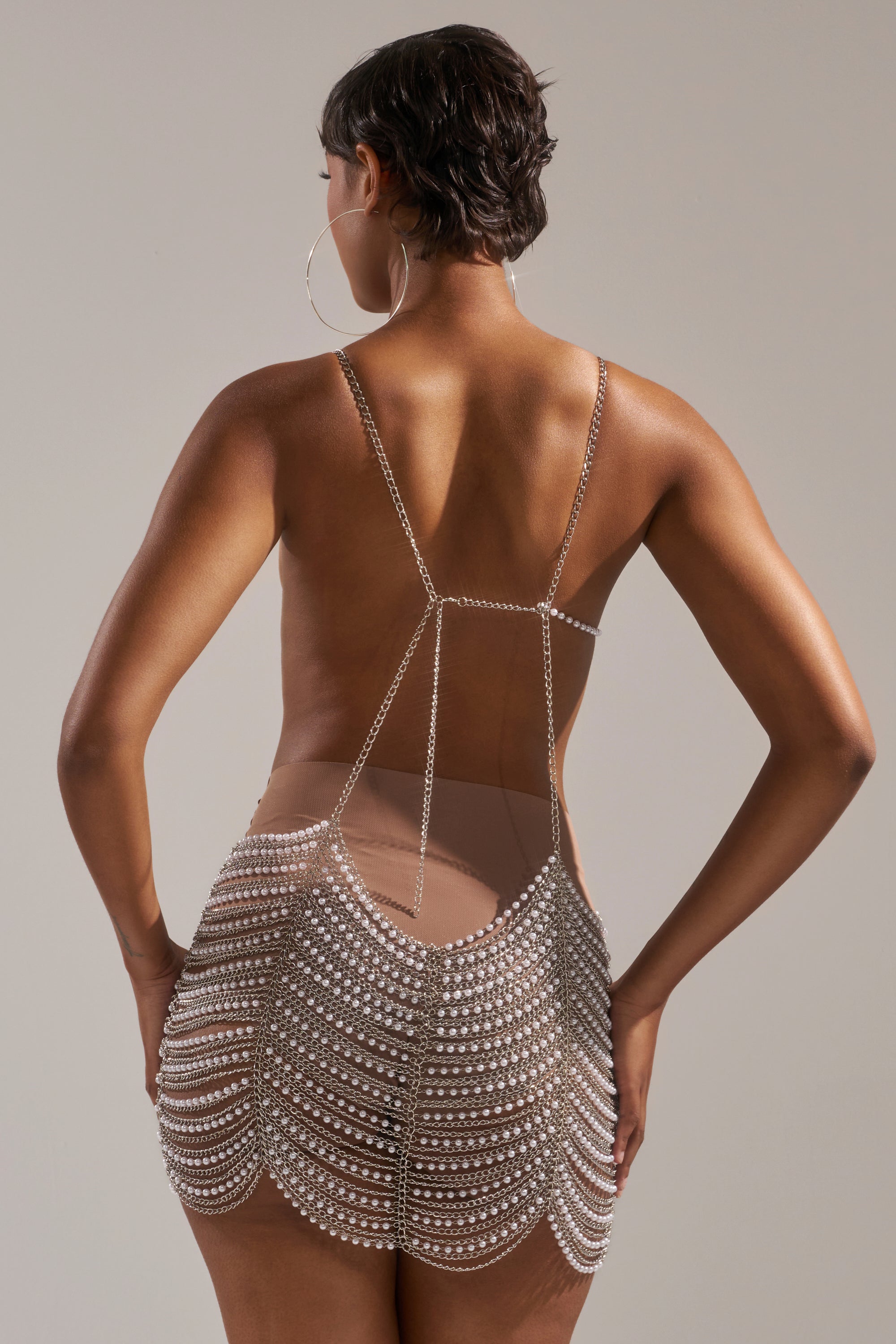 A person with short dark hair faces away, wearing the ITS GIVING DRAMA CHAIN MINI DRESS—a curve-hugging mini adorned with sparkling silver chains and scalloped beadwork over the hips, finished with large hoop earrings.