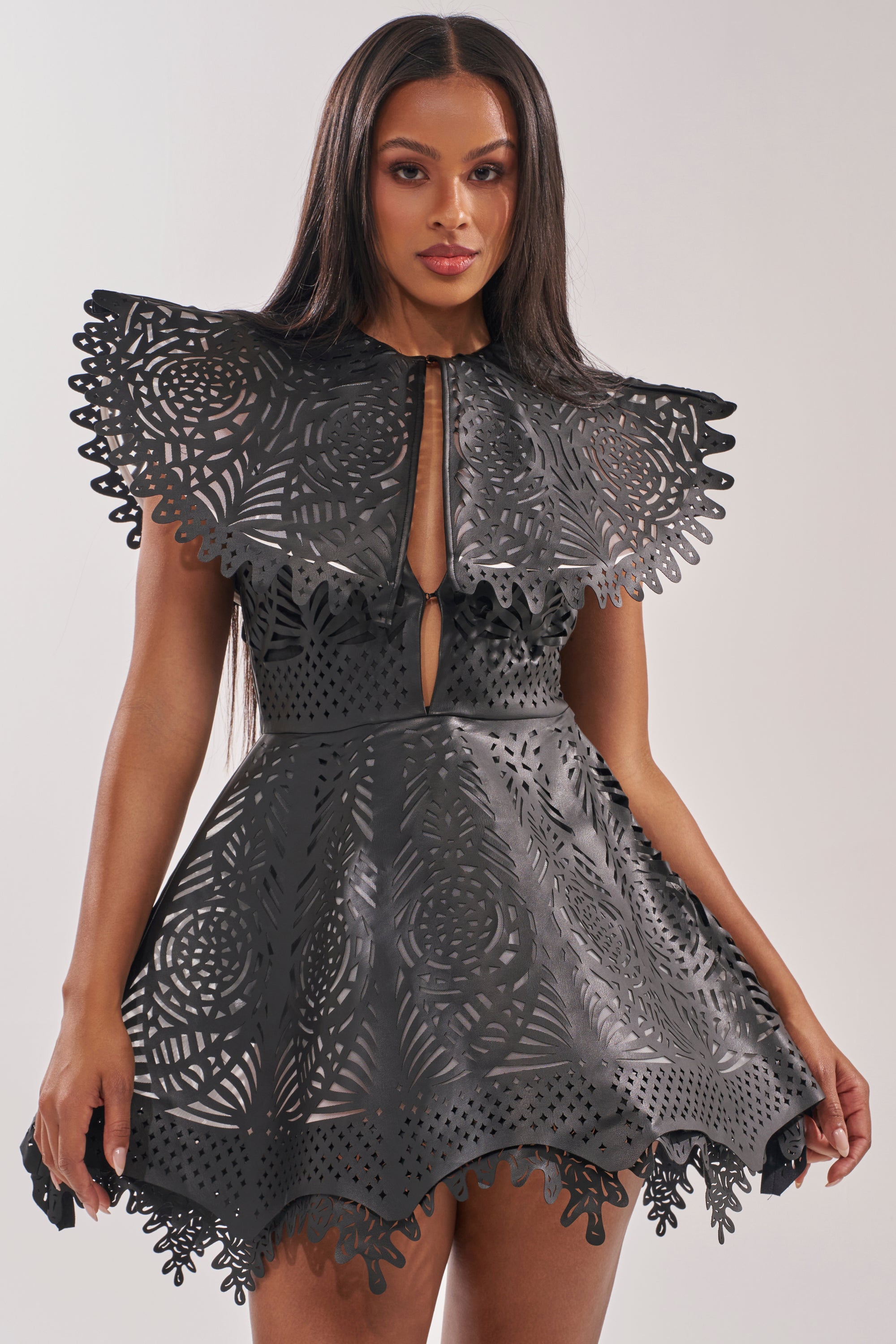 A woman wears the URBAN EDGE MINI COLLARED DRESS—a black faux leather mini with laser-cut patterns, scalloped edges, and bold structured shoulders—posing against a light background for a striking cocktail look. 0
