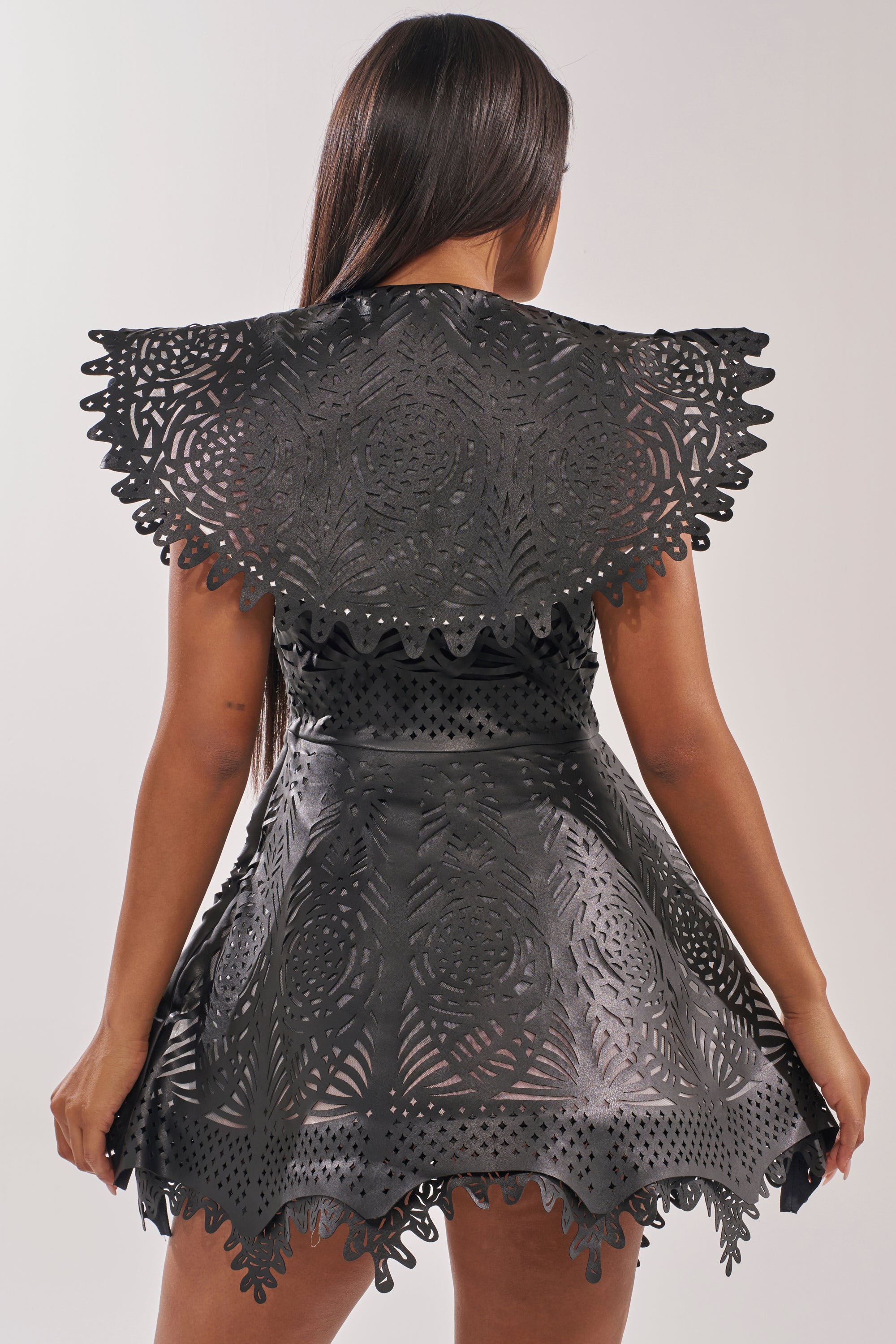 A woman with long dark hair faces away, wearing the URBAN EDGE MINI COLLARED DRESS—a black faux leather, fit-and-flare mini dress with laser-cut symmetrical patterns, scalloped shoulders, and a textured flared skirt. 3