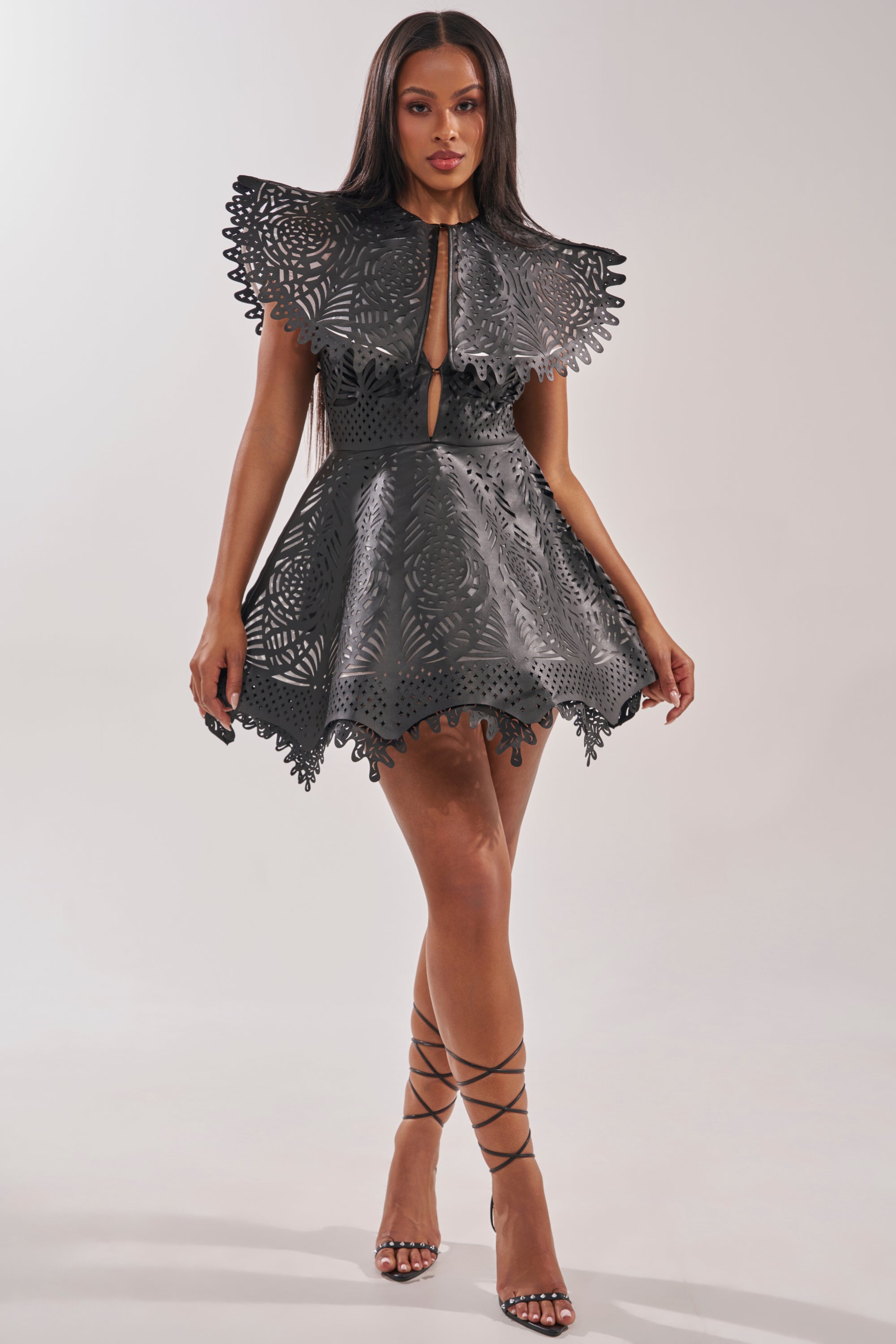 A woman wears the URBAN EDGE MINI COLLARED DRESS, a black faux leather mini with structured laser-cut patterns and a cape-style collar. She pairs it with strappy high-heeled sandals for an elegant cocktail party look. 5
