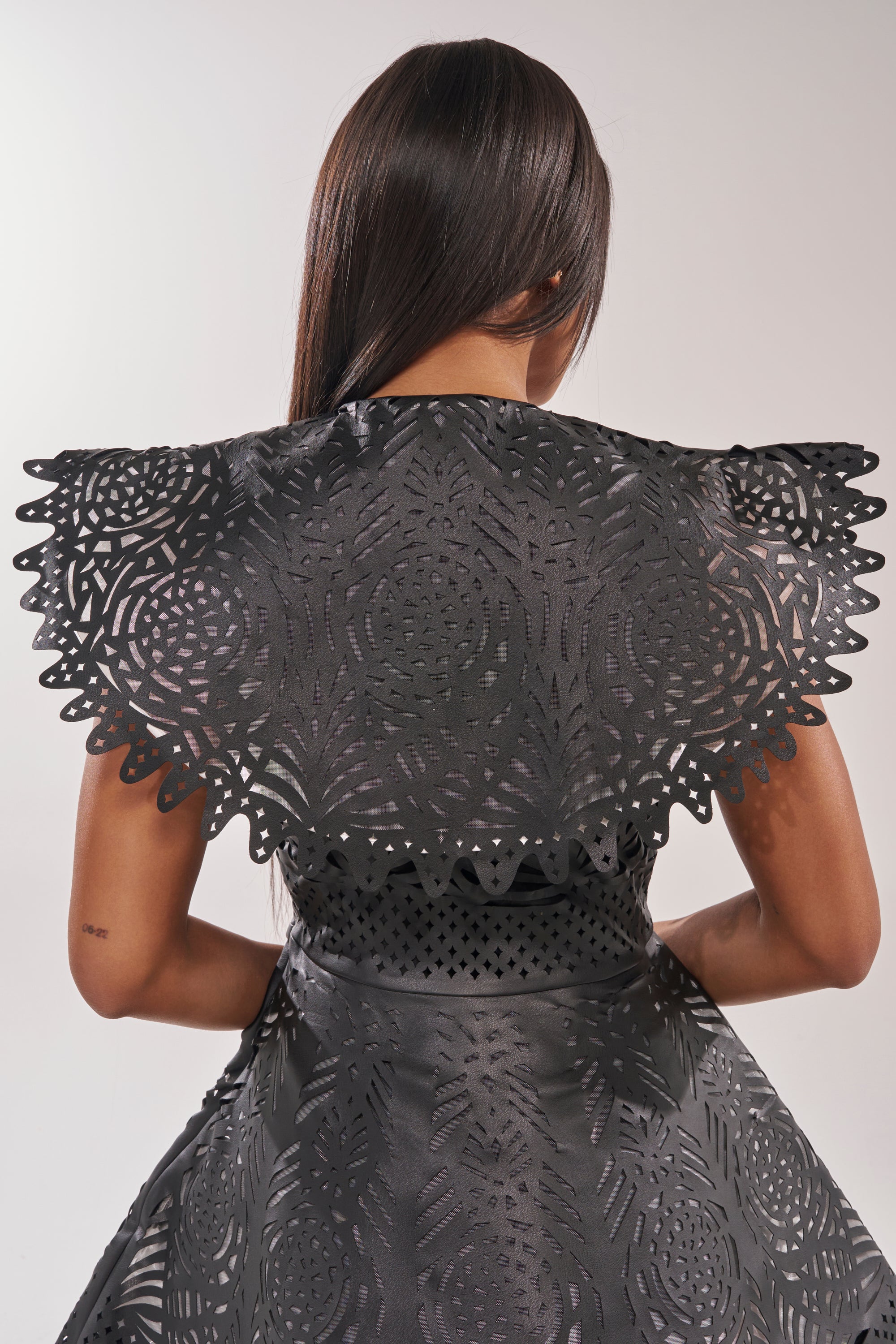 A woman with long straight hair is seen from behind in a black URBAN EDGE MINI COLLARED DRESS featuring laser-cut patterns and a dramatic collar, set against a plain light background. 6