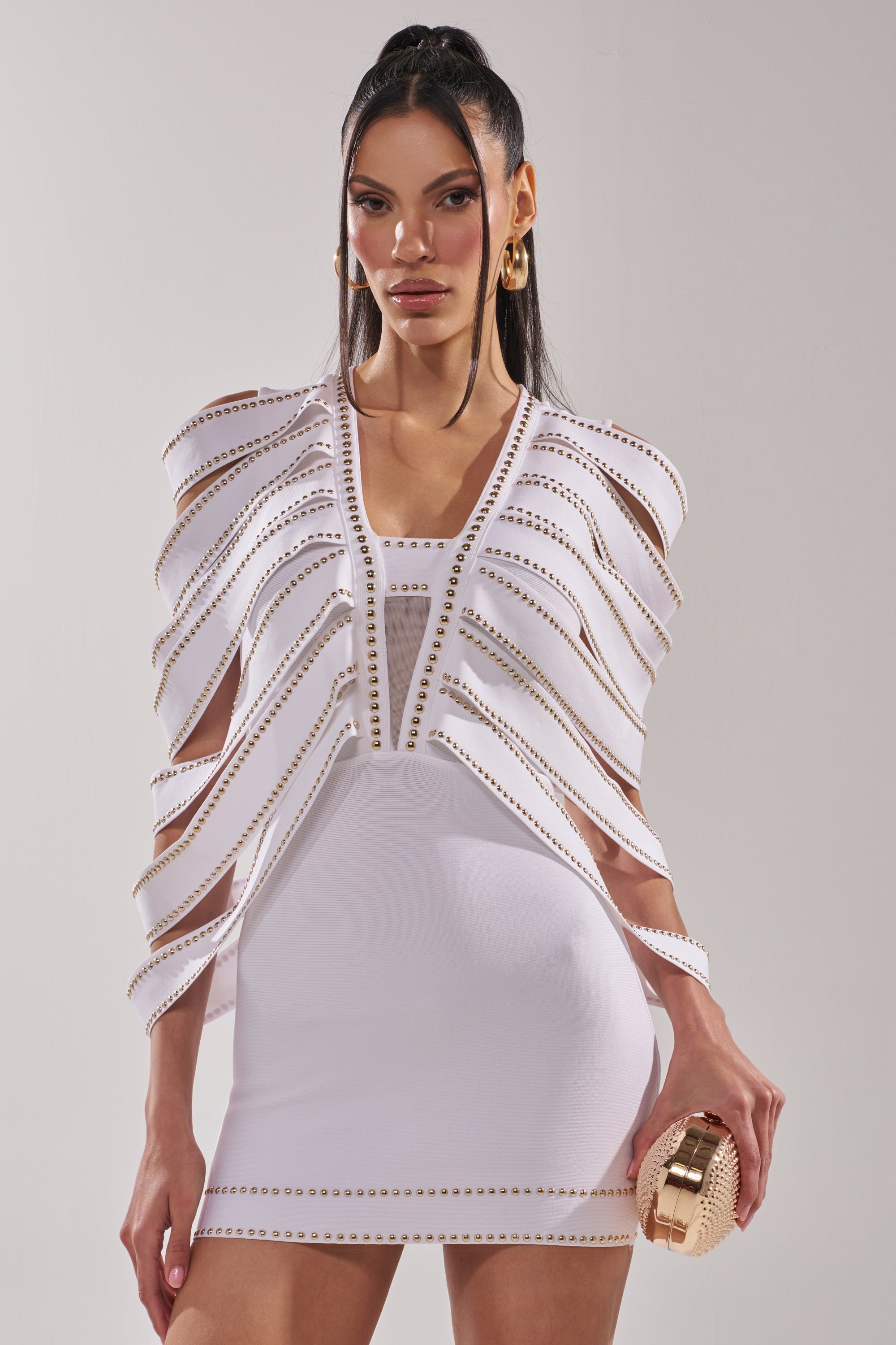 A woman with long dark hair wears the ANGEL WINGS MINI DRESS, a white mini dress with gold accents and a structured cape detail. She pairs it with a gold clutch and hoop earrings, standing out against a plain background. 0