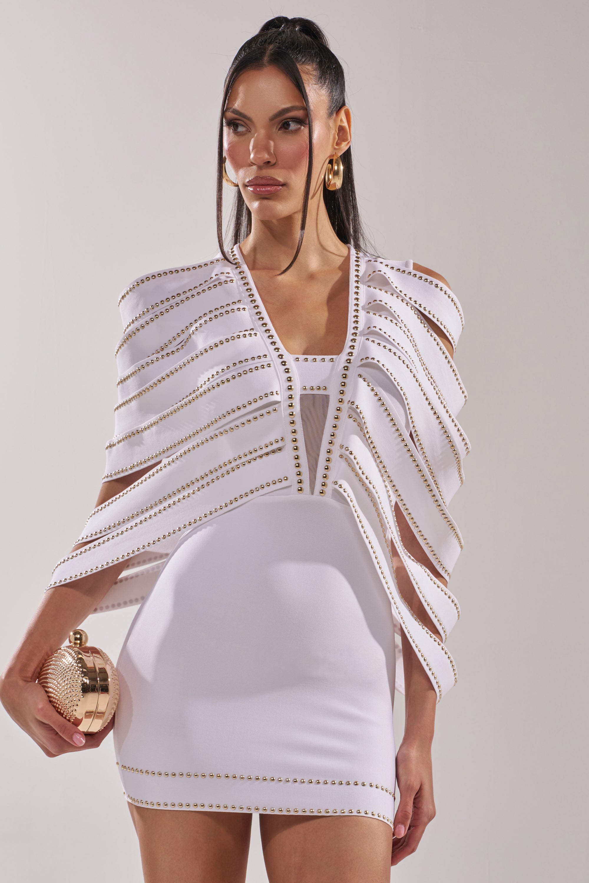 A woman wears the ANGEL WINGS MINI DRESS, a form-fitting white mini dress with gold studs and cape-like panels, styled with gold hoop earrings, a high ponytail, and a round gold clutch. 1