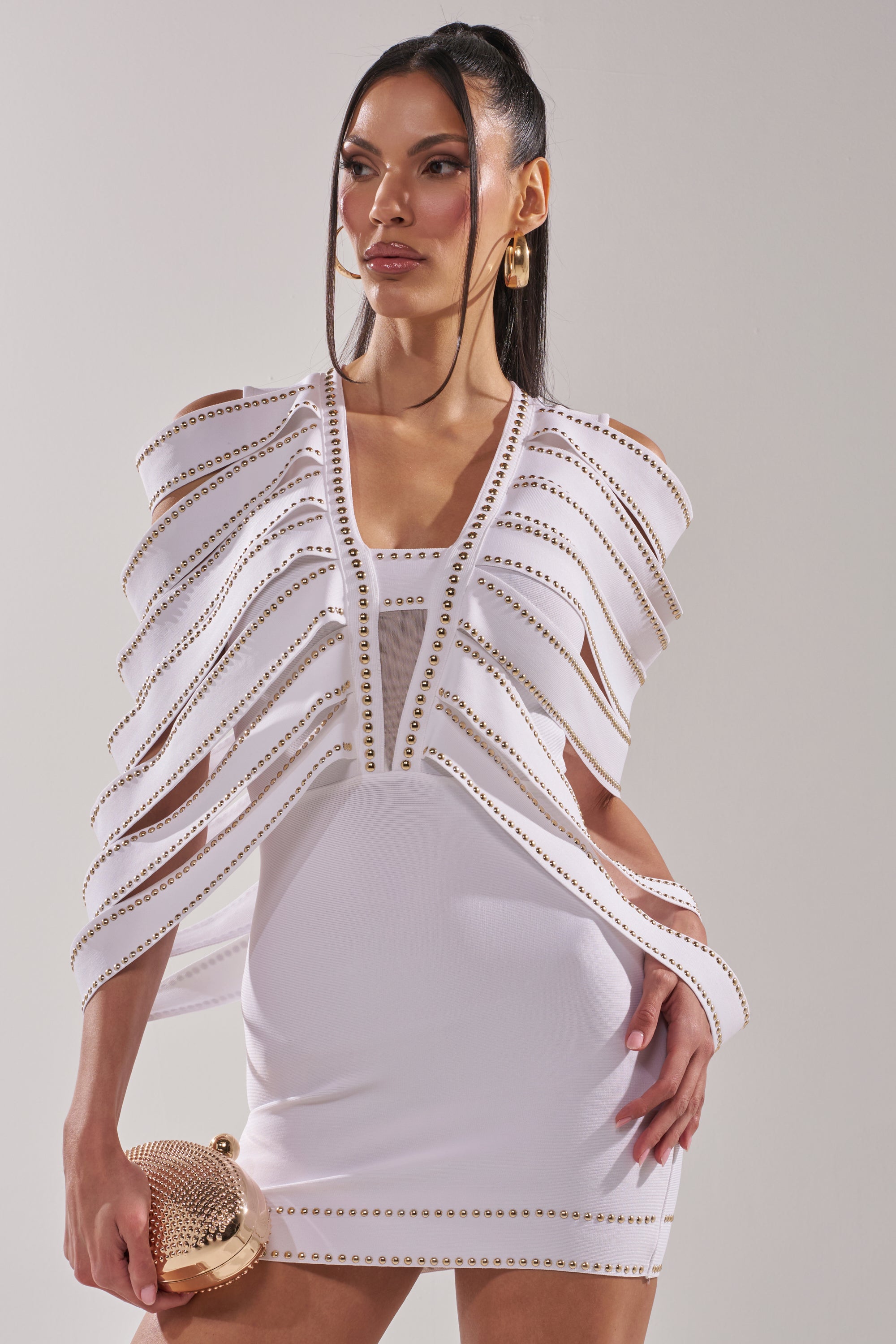 Wearing the ANGEL WINGS MINI DRESS—a fitted white mini with winged layers and gold studs—a woman holds a gold clutch, her long straight hair and hoop earrings completing the bold look as she poses confidently against a plain background. 2
