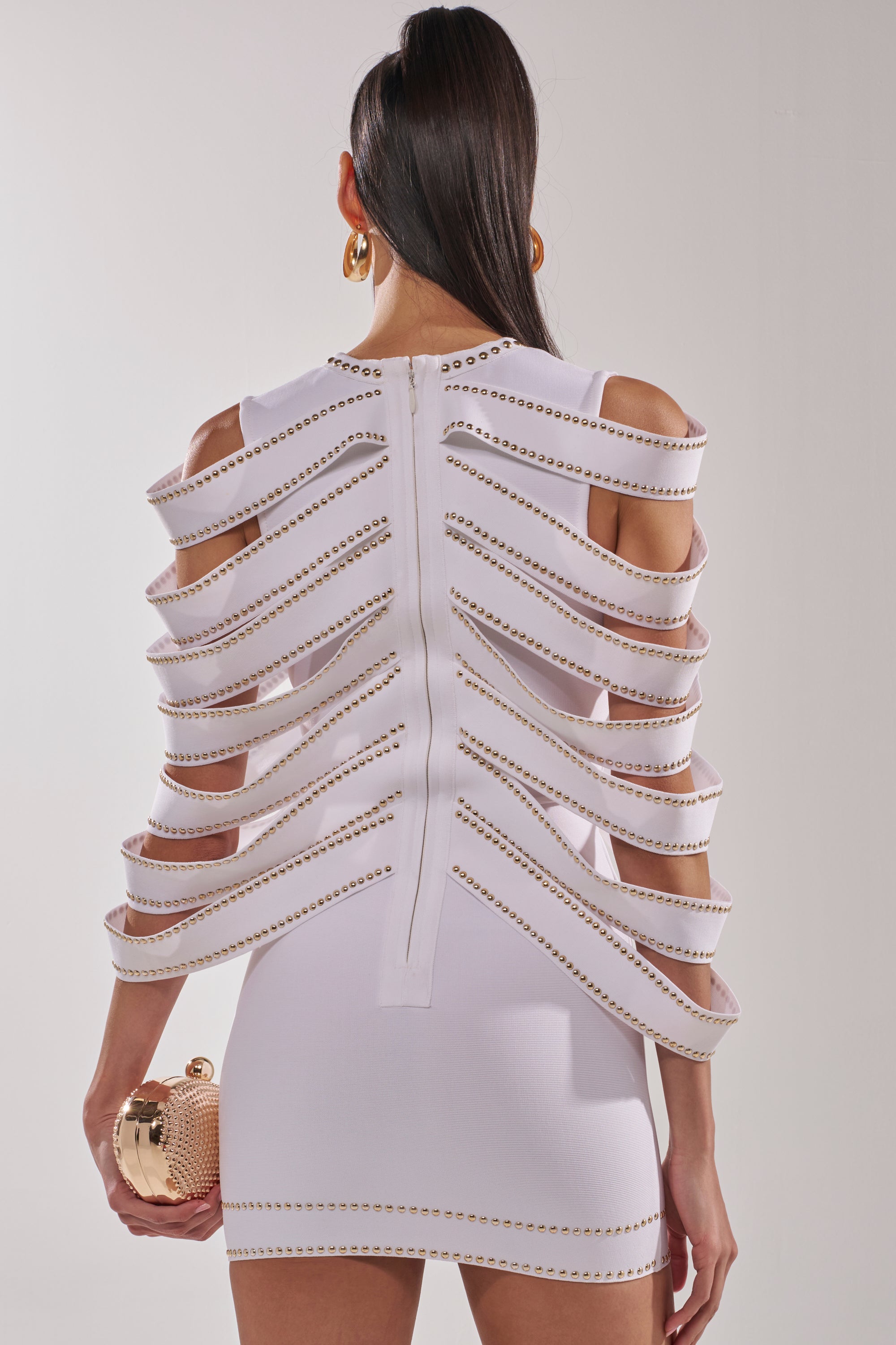 Wearing the ANGEL WINGS MINI DRESS, a white bodycon mini with horizontal cutouts and metallic studs, she pairs it with a round gold clutch and large hoop earrings. Her straight, sleek hair completes this chic look. 4