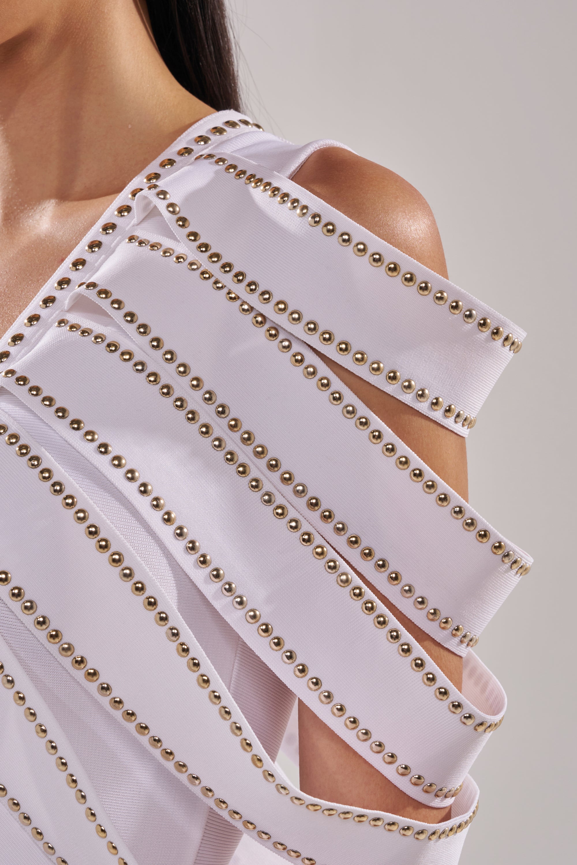 Close-up of a woman wearing the ANGEL WINGS MINI DRESS, a white mini dress with multiple horizontal bands and gold studs, creating a layered and structured effect for a modern, studded cape-style look. 8