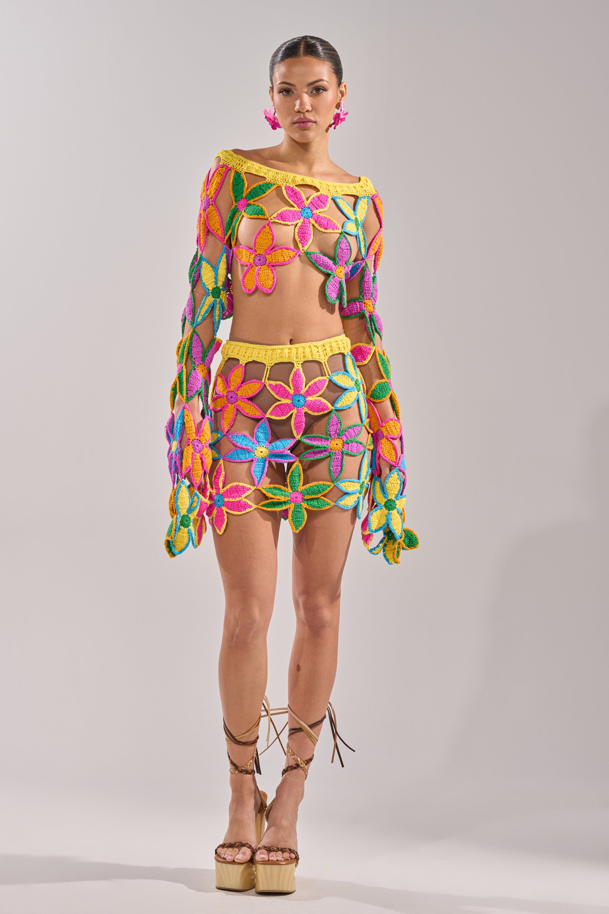A woman wears the FLORAL FIELDS CROCHET OFF THE SHOULDER SWEATER, featuring sheer multicolored floral crochet and flared sleeves. She pairs it with pink earrings, lace-up platform sandals, and a tied-back hairstyle. 11