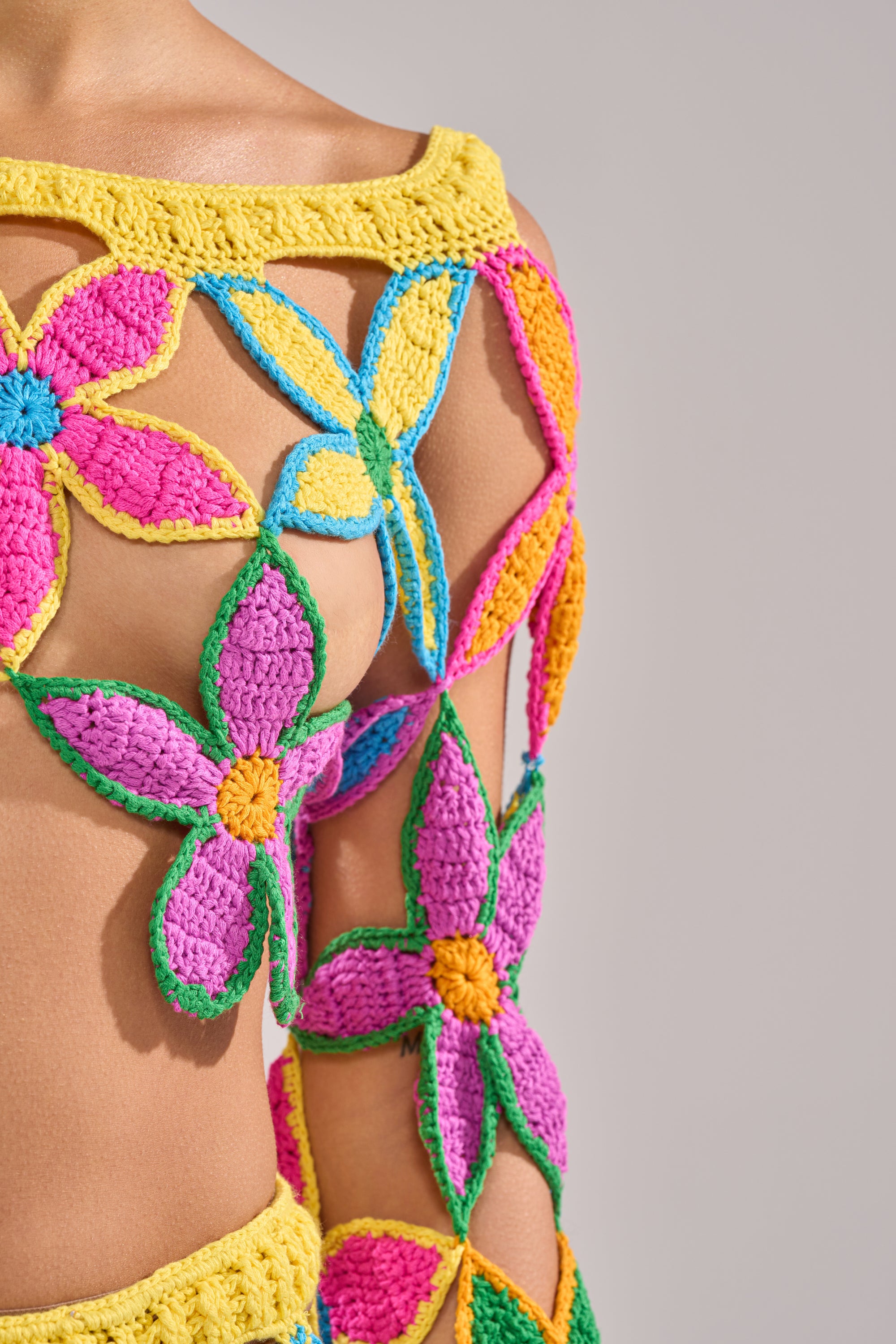A close-up of a person wearing the FLORAL FIELDS CROCHET OFF THE SHOULDER SWEATER, featuring vibrant open flower patterns in yellow, pink, purple, blue, and orange yarn that reveal skin underneath. 12