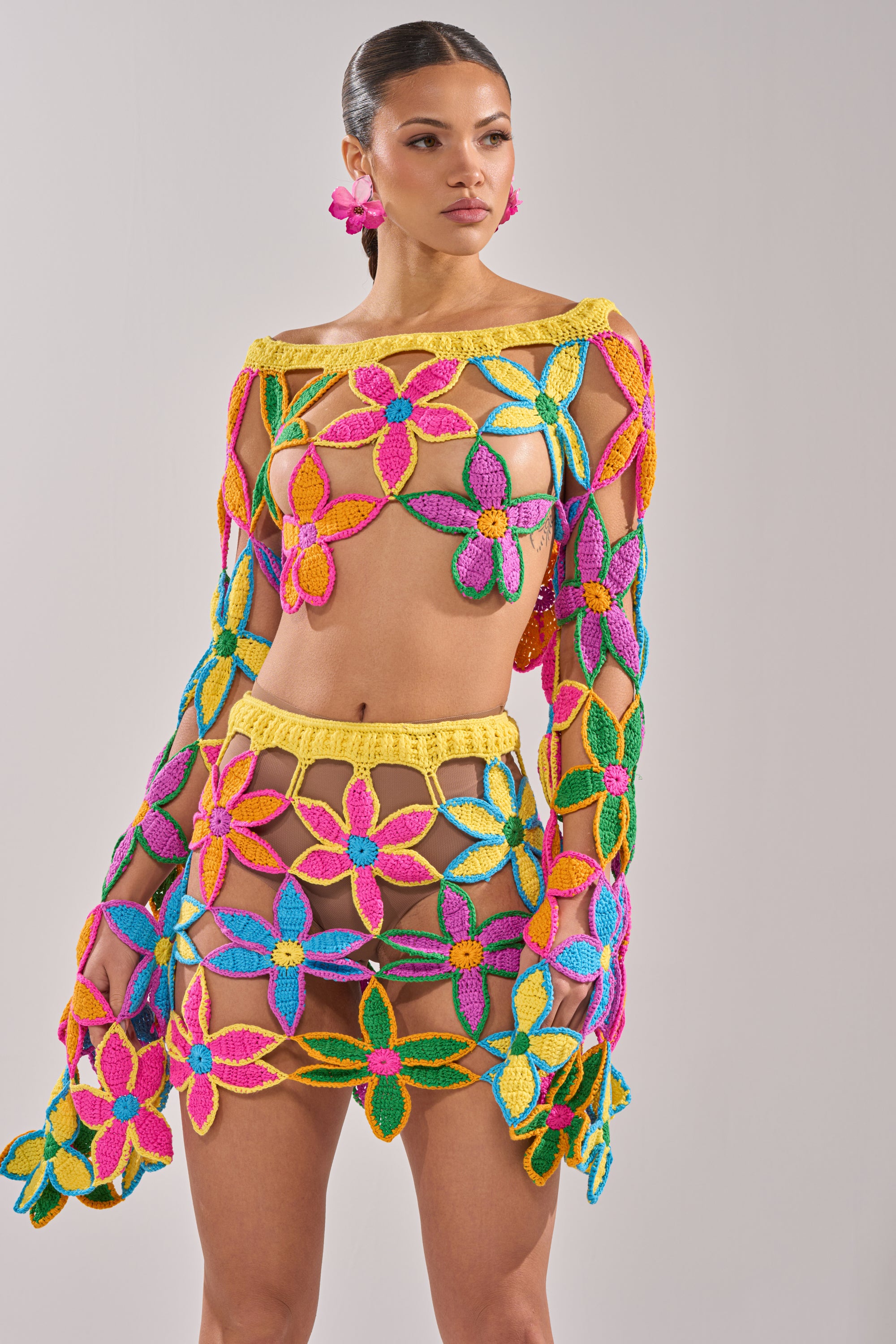 A woman wears the FLORAL FIELDS CROCHET OFF THE SHOULDER SWEATER, a colorful sheer knit with yellow, pink, blue, and green floral patterns. Paired with matching flower earrings, it’s a vibrant choice for summer festivals against a plain background. 3