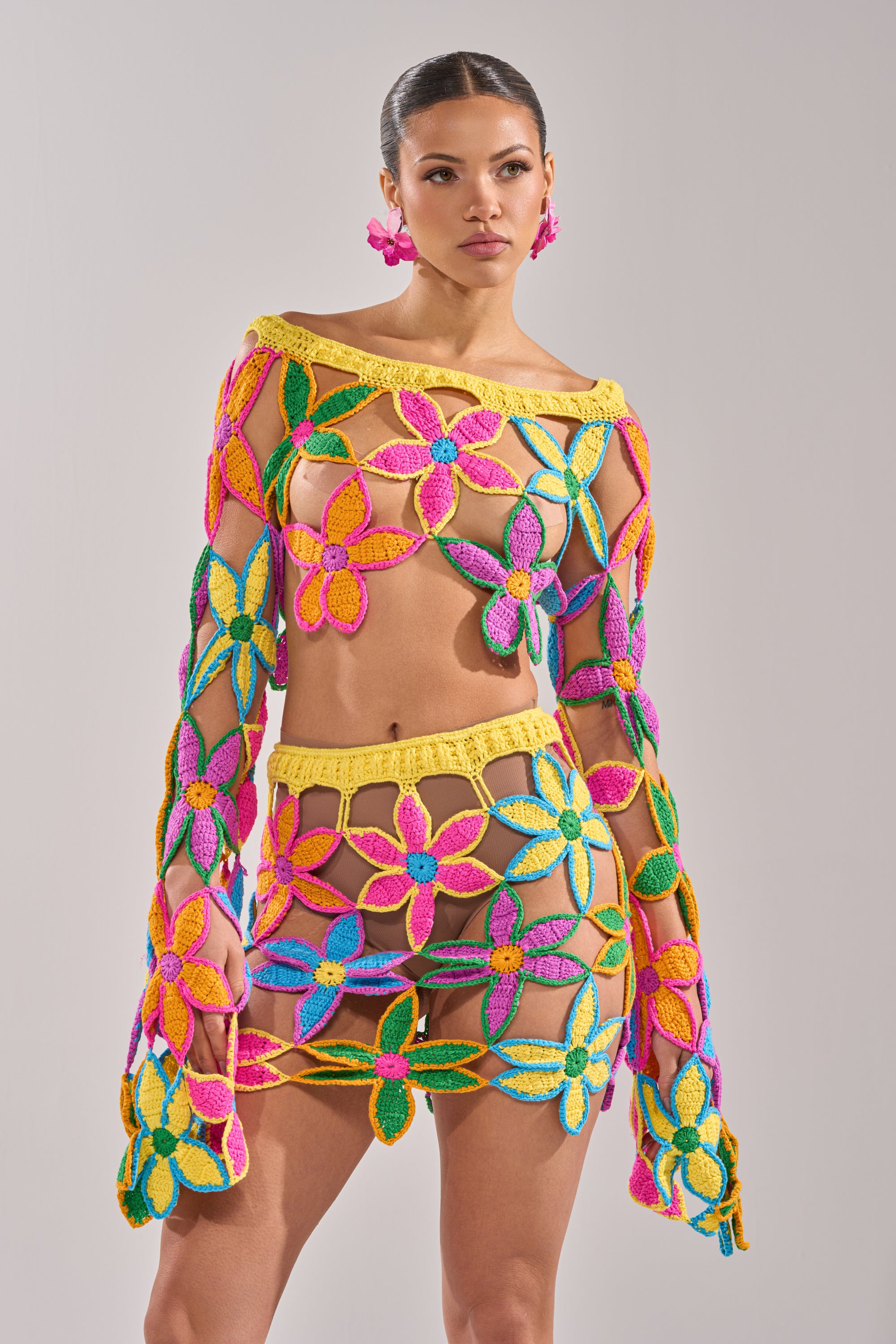 A woman models the FLORAL FIELDS CROCHET OFF THE SHOULDER SWEATER, featuring large open flowers in pink, yellow, green, and blue, paired with matching floral earrings against a plain light background. 5