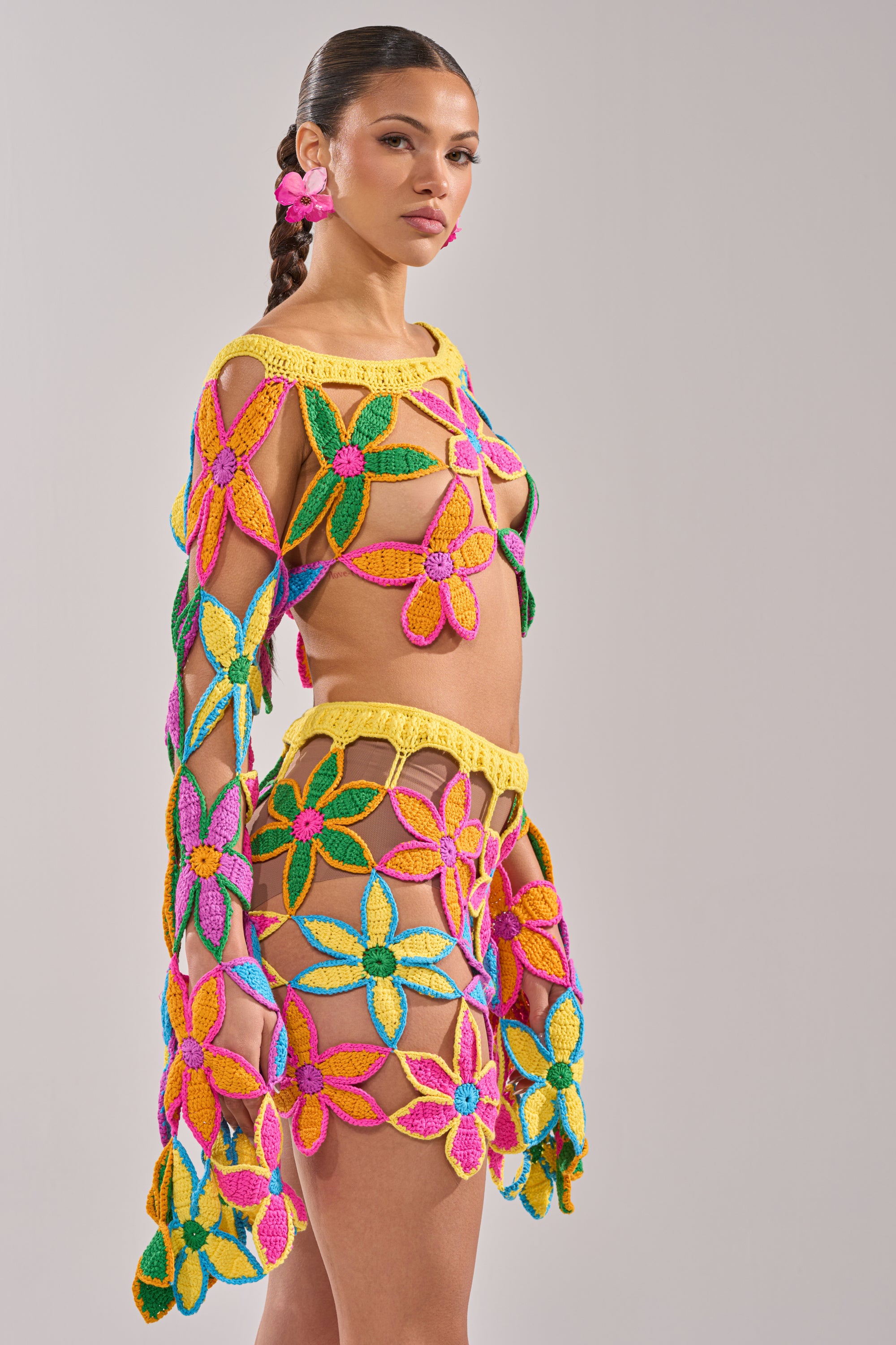 A woman with a braid and pink flower earrings wears the FLORAL FIELDS CROCHET OFF THE SHOULDER SWEATER, featuring large yellow, pink, green, and blue floral patterns—an eye-catching choice for summer parties. 8