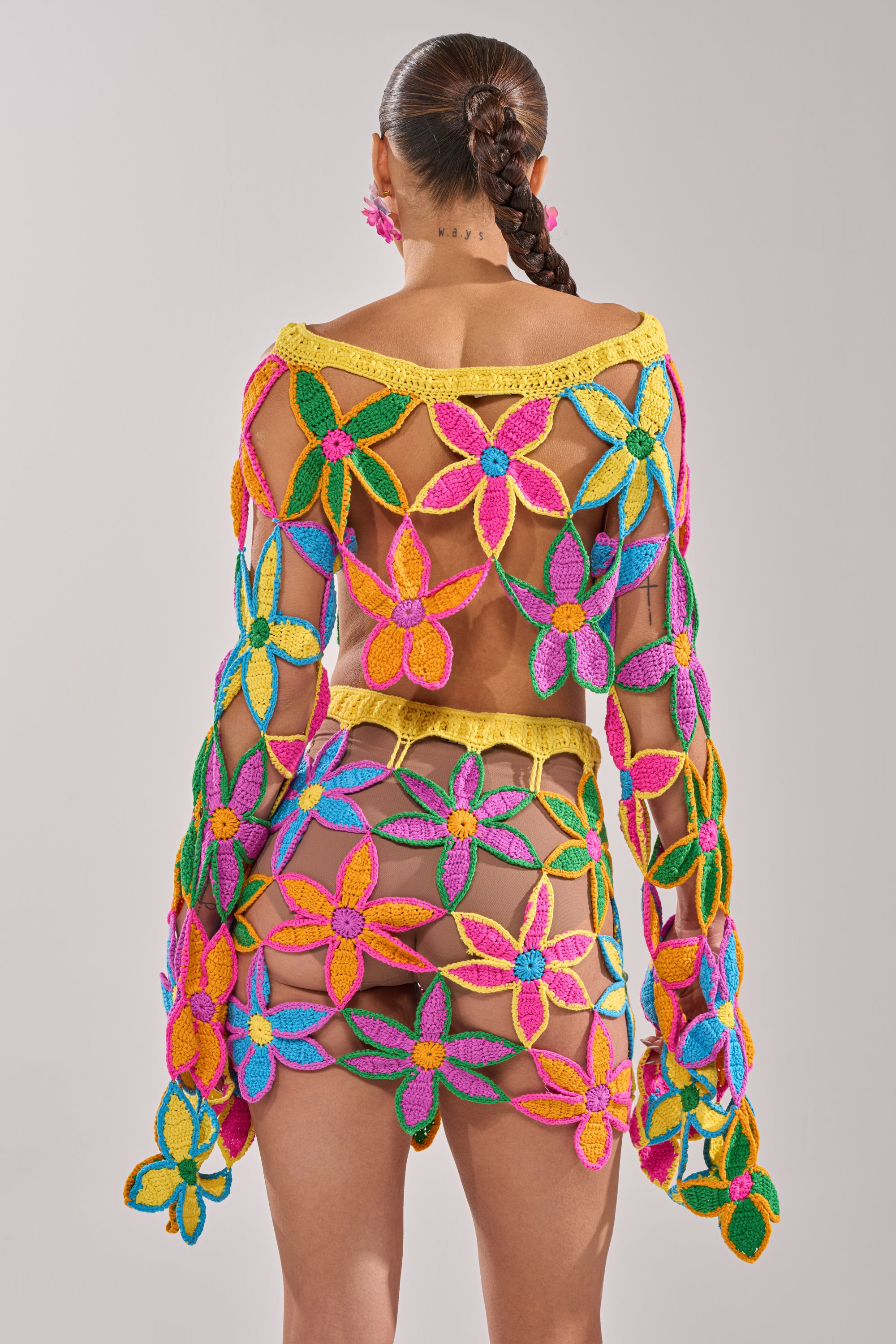 A person with a braided ponytail wears the FLORAL FIELDS CROCHET OFF THE SHOULDER SWEATER, featuring vibrant multicolored floral motifs and an open-back design, against a plain light gray background. 9