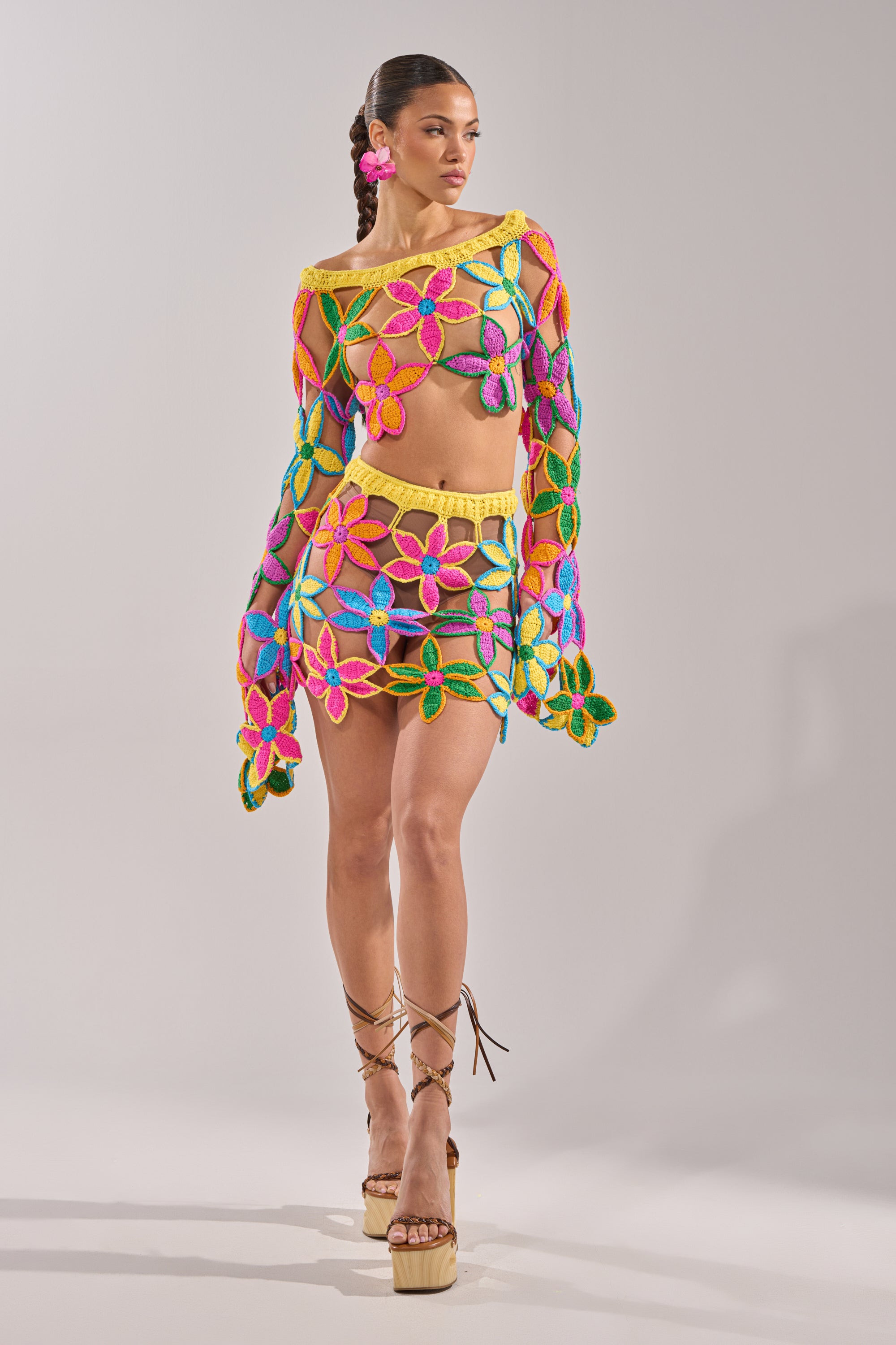 A woman poses in the FLORAL FIELDS CROCHET OFF THE SHOULDER SWEATER, featuring vibrant sheer floral crochet and colorful flower patterns—an eye-catching summer party top, styled with a matching mini skirt and playful pink accessories. 10