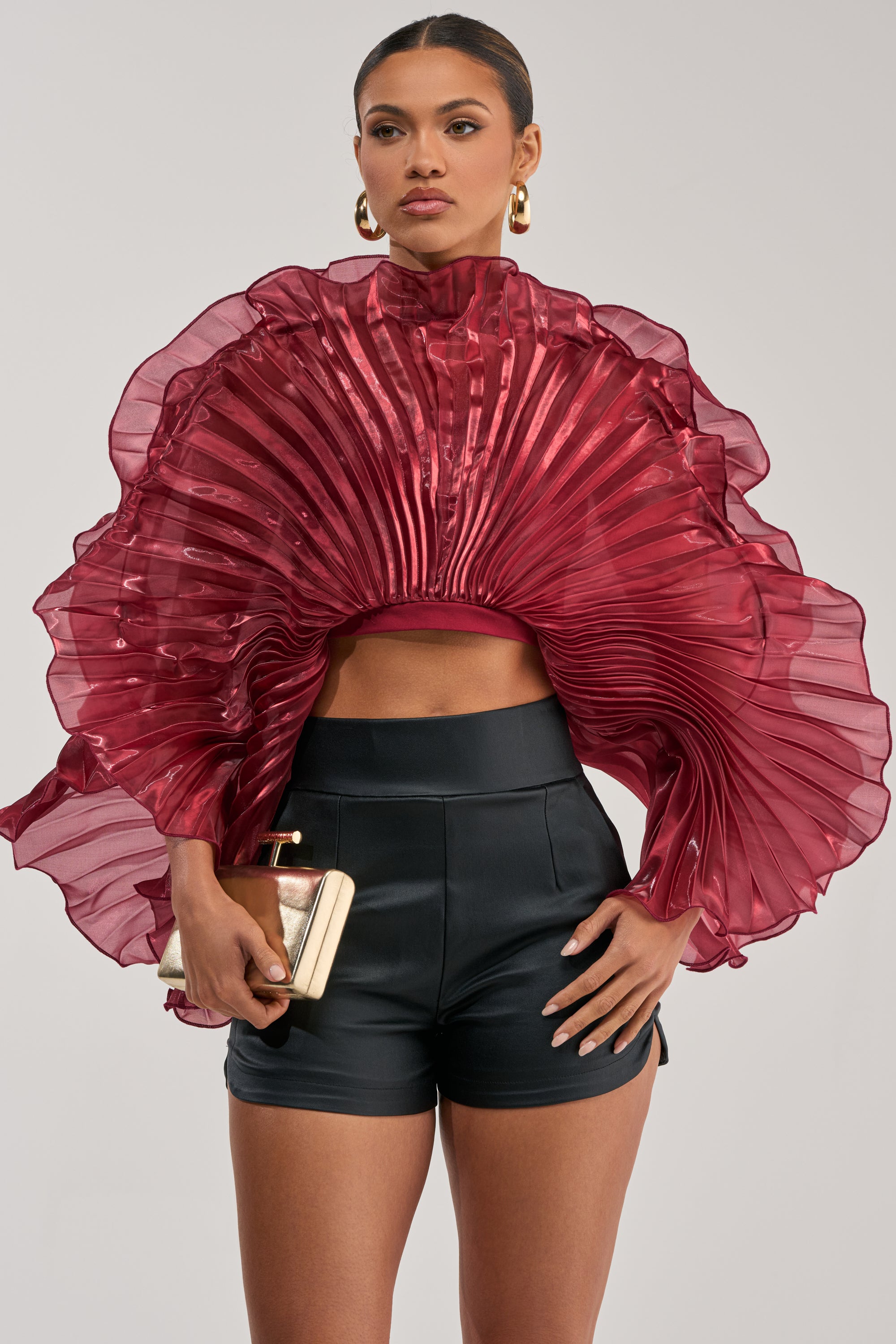 A woman wears the SHINING DOWN ON YOU BLOUSE IN BURGUNDY with dramatic pleated ruffles, paired with high-waisted black shorts, gold hoop earrings, and a gold clutch against a plain background.