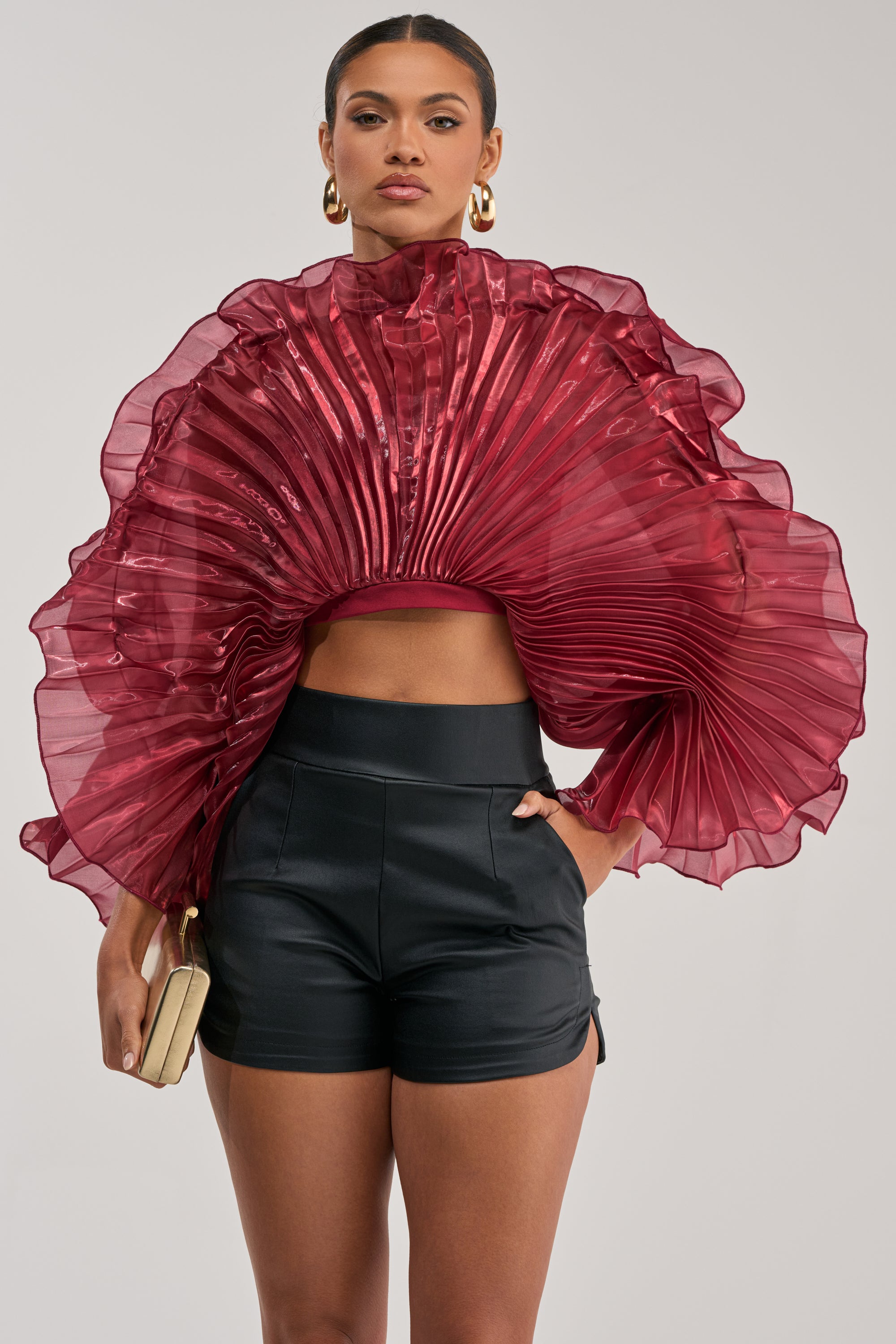 A woman in gold hoop earrings, black high-waisted shorts, and a small beige clutch wears the SHINING DOWN ON YOU BLOUSE IN BURGUNDY with dramatic pleated ruffles, standing against a plain light background.
