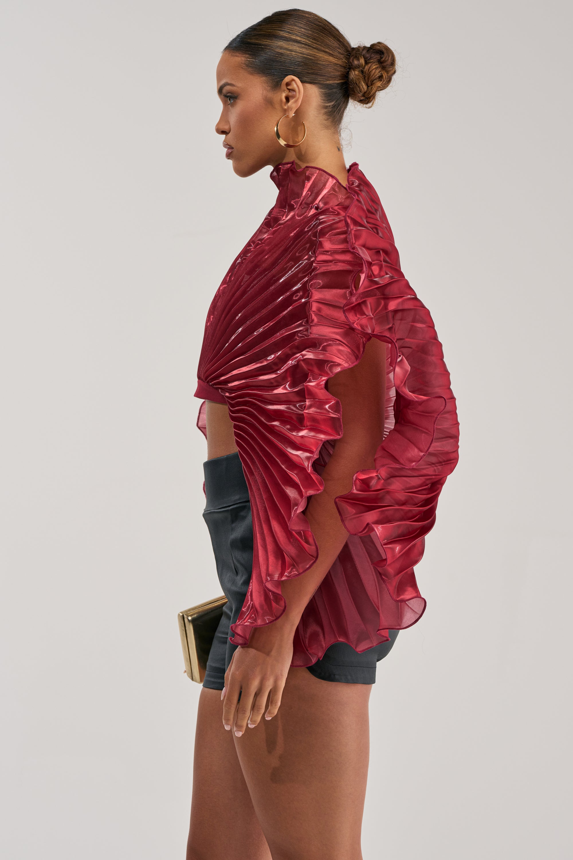 A woman in profile holds a beige clutch, wearing the SHINING DOWN ON YOU BLOUSE IN BURGUNDY with pleated ruffles and a high collar, paired with black shorts and gold hoop earrings against a light background.