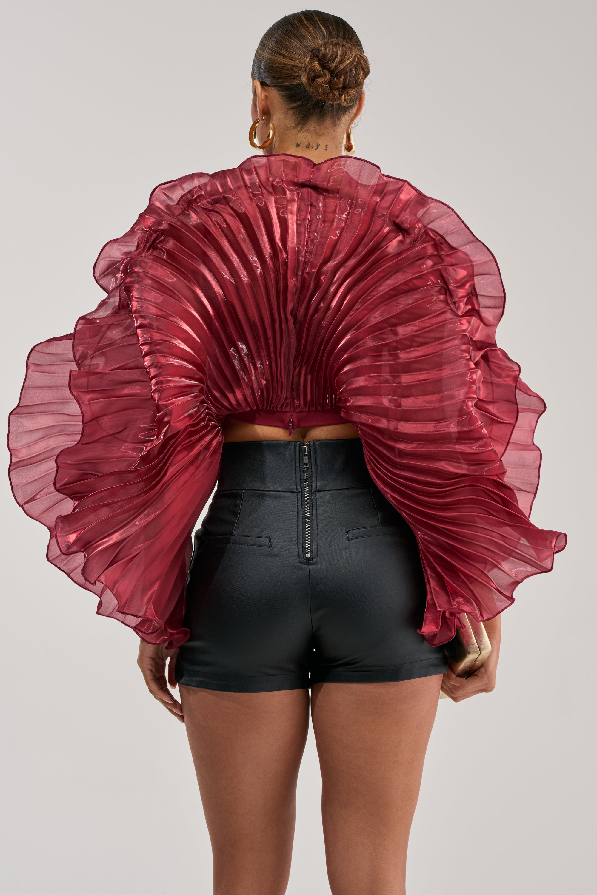 A woman faces away, wearing the SHINING DOWN ON YOU BLOUSE IN BURGUNDY—an avant-garde sheer, pleated top with ruffled sleeves—and high-waisted black faux leather shorts. Her sleek bun and gold hoop earrings complete the look.